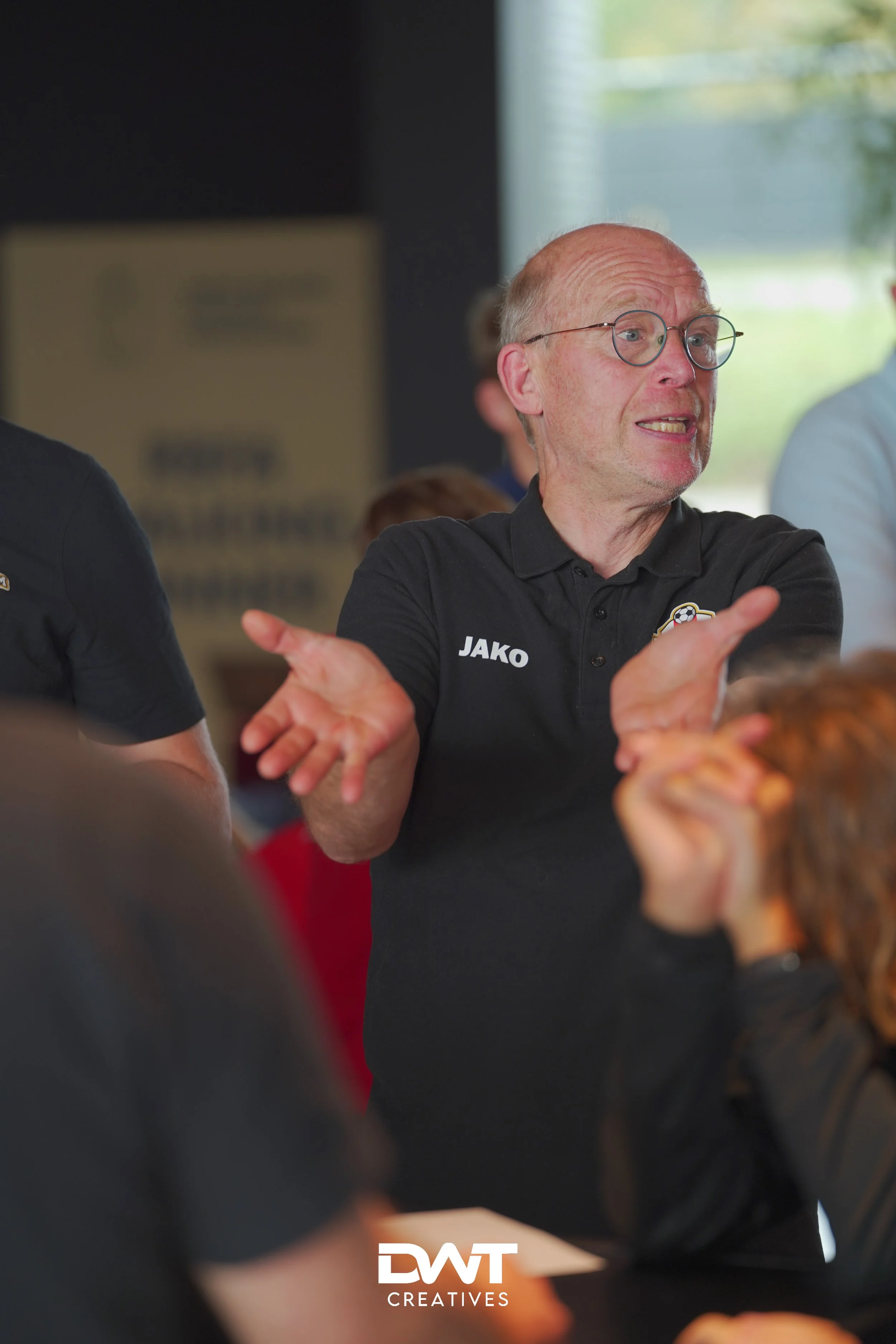 Een persoon met een bril en een zwart poloshirt gebaart met zijn armen in een binnenomgeving. Op de achtergrond zijn onscherpe mensen zichtbaar.