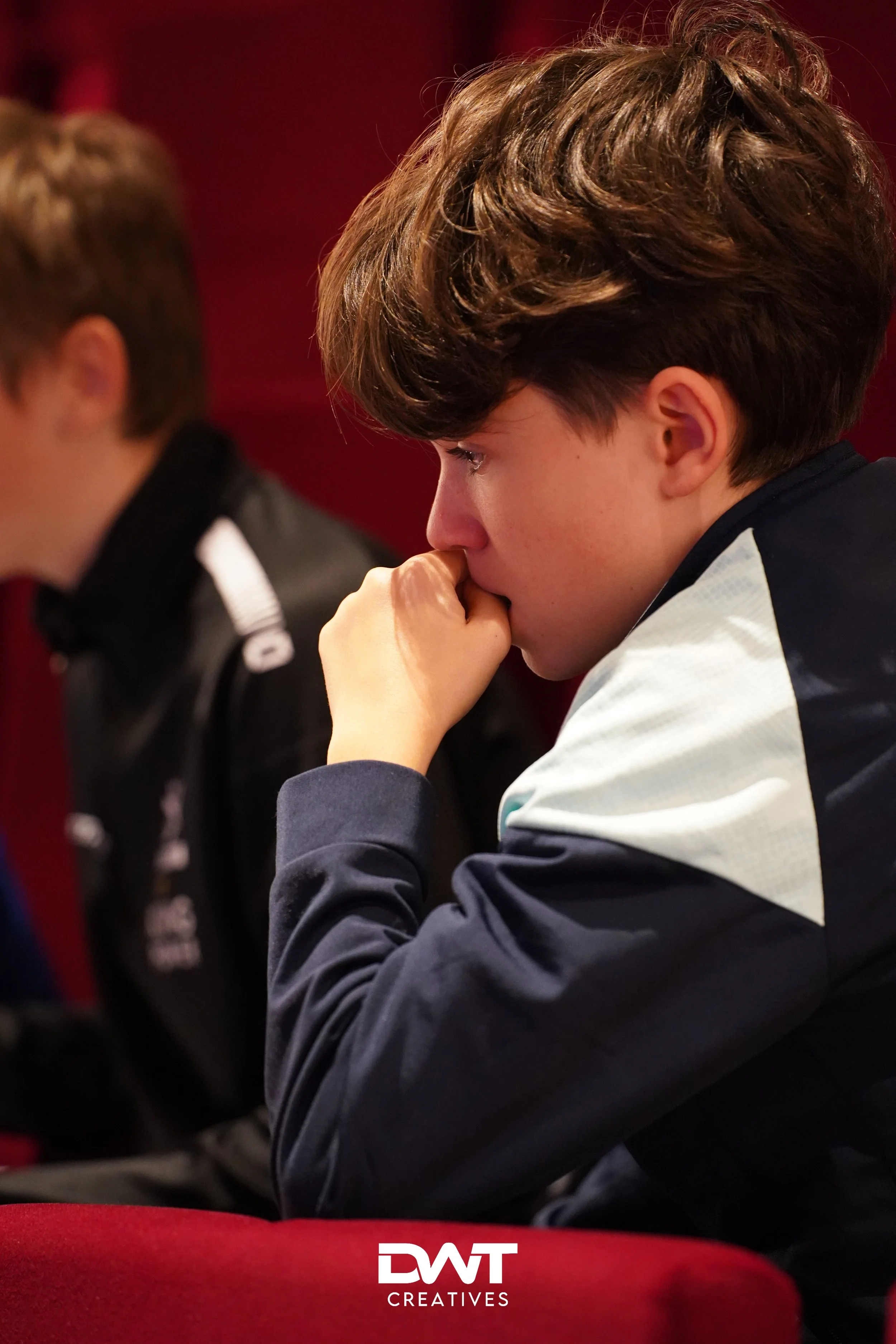 Twee jongeren zitten naast elkaar in een rode ruimte. De jongen op de voorgrond leunt met zijn hand tegen zijn kin, terwijl hij geconcentreerd kijkt. Het logo "DWT Creatives" staat rechtsonder in de afbeelding.