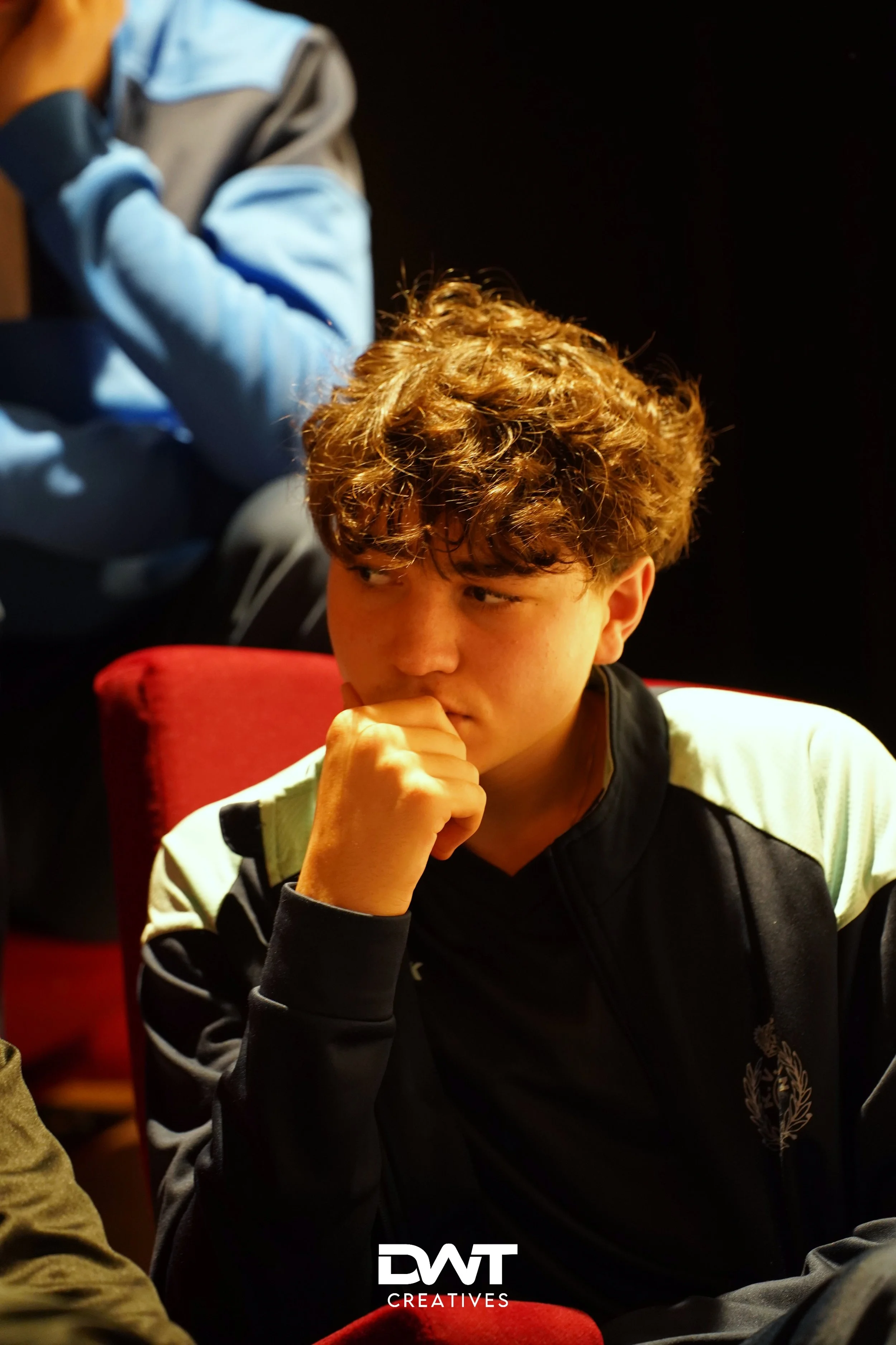 Een jongen met krullend haar zit op een rode stoel en houdt zijn hand aan zijn kin in een nadenkende houding.