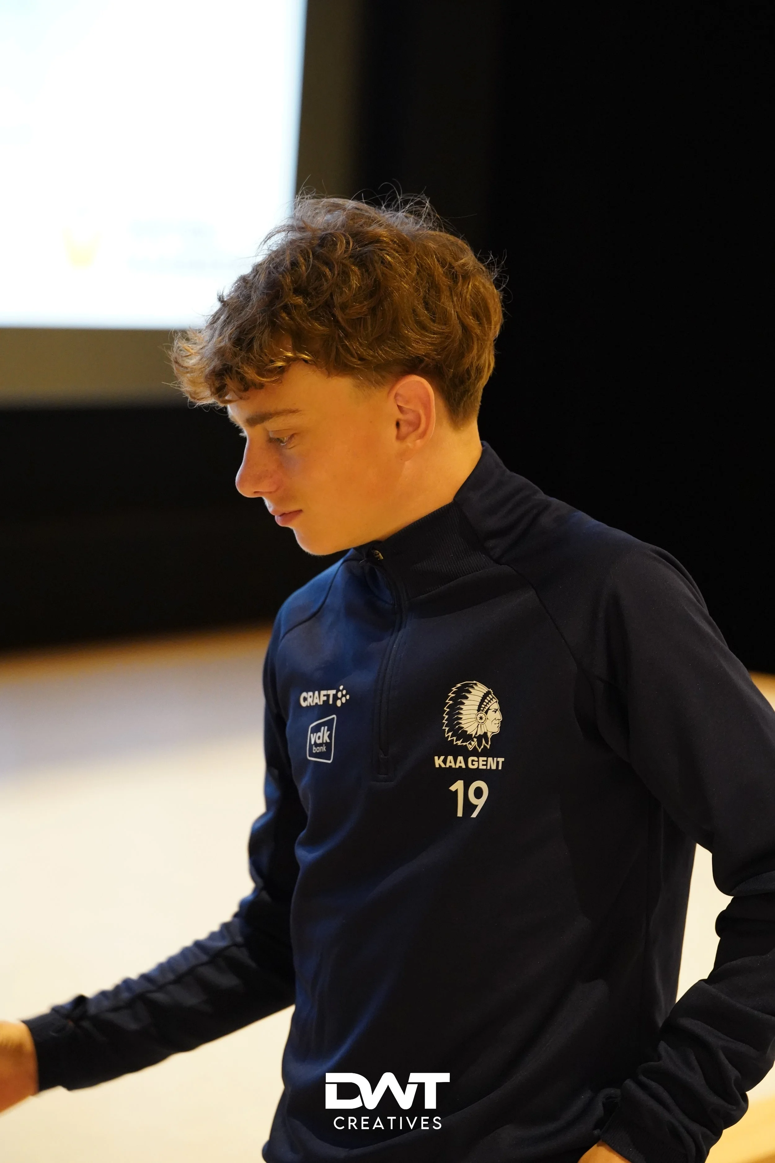 Jonge persoon in een donkerblauw KAA Gent-jack met een logo en nummer. De achtergrond is onscherp. Het jack heeft ook het merk Craft en een vdk bank logo.