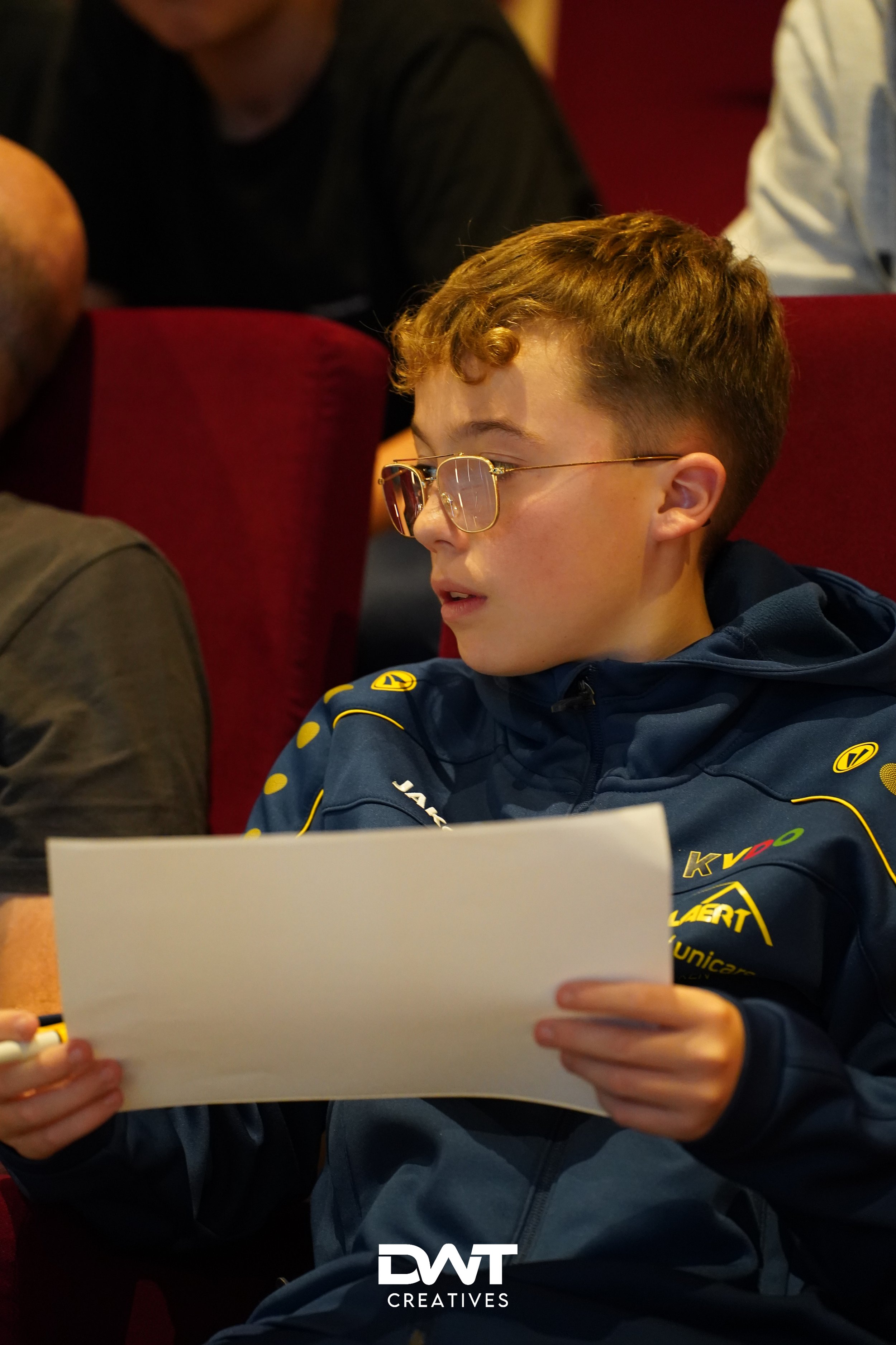 Jongen met bril leest een papier, zittend in een rode stoel, geconcentreerd kijkend, gekleed in een sportief blauw jasje met gele details.