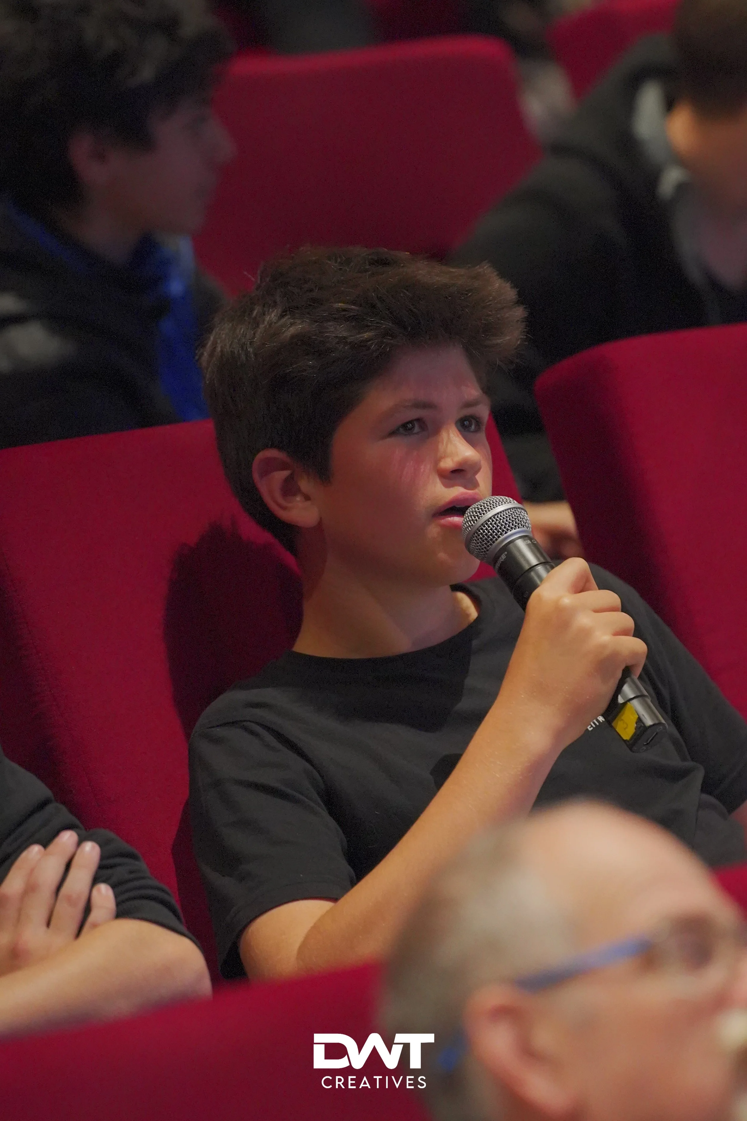 Jongen in zwart T-shirt met microfoon, zittend in rode stoel tijdens evenement.