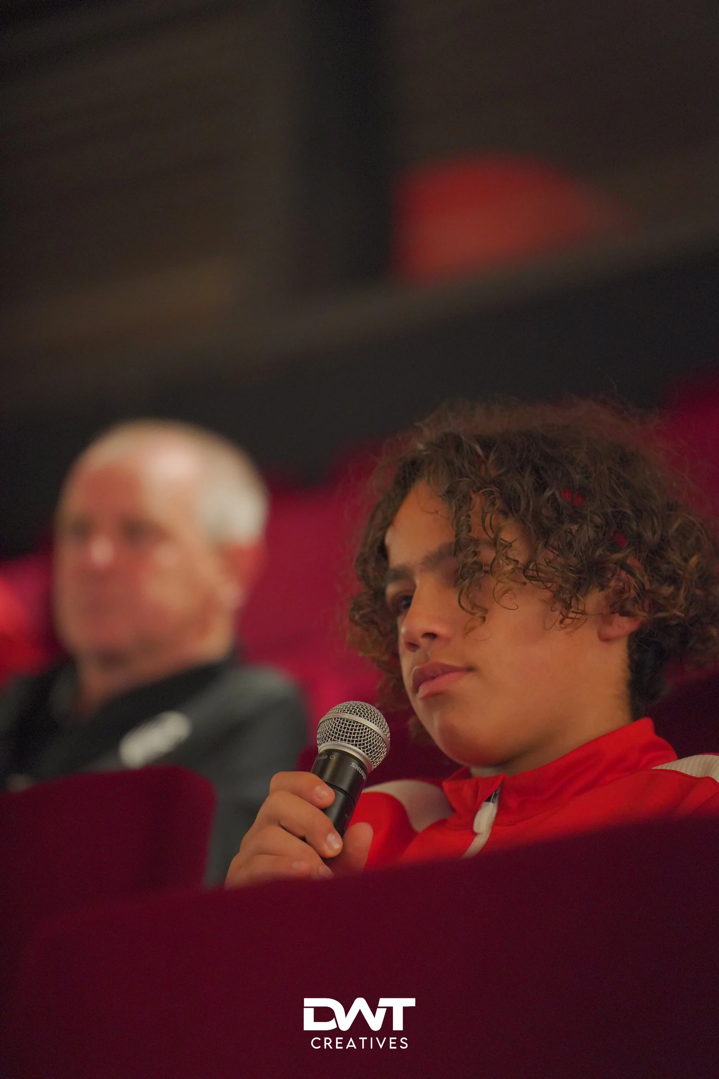 Een jongen met krullend haar houdt een microfoon vast, zittend in een bioscoop- of theaterstoel. Er is een oudere man vaag zichtbaar op de achtergrond. Het logo "DWT Creatives" staat onderaan de afbeelding.