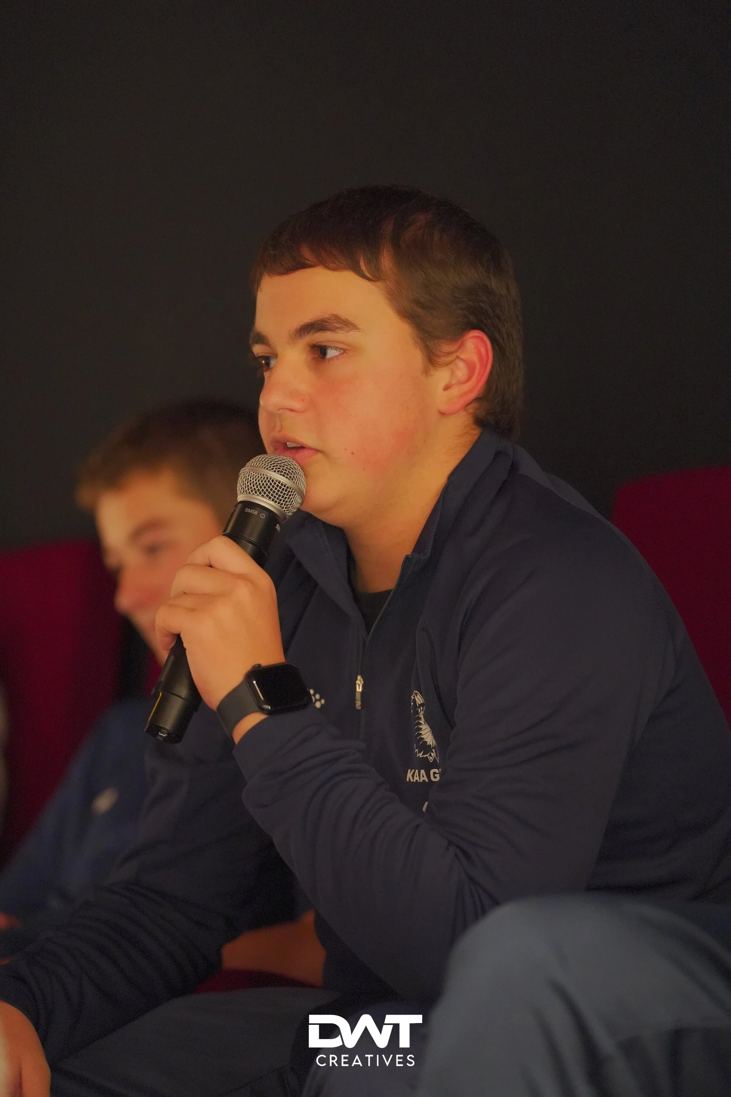 Jonge man met microfoon in blauwe jas, spreekt tijdens een evenement.