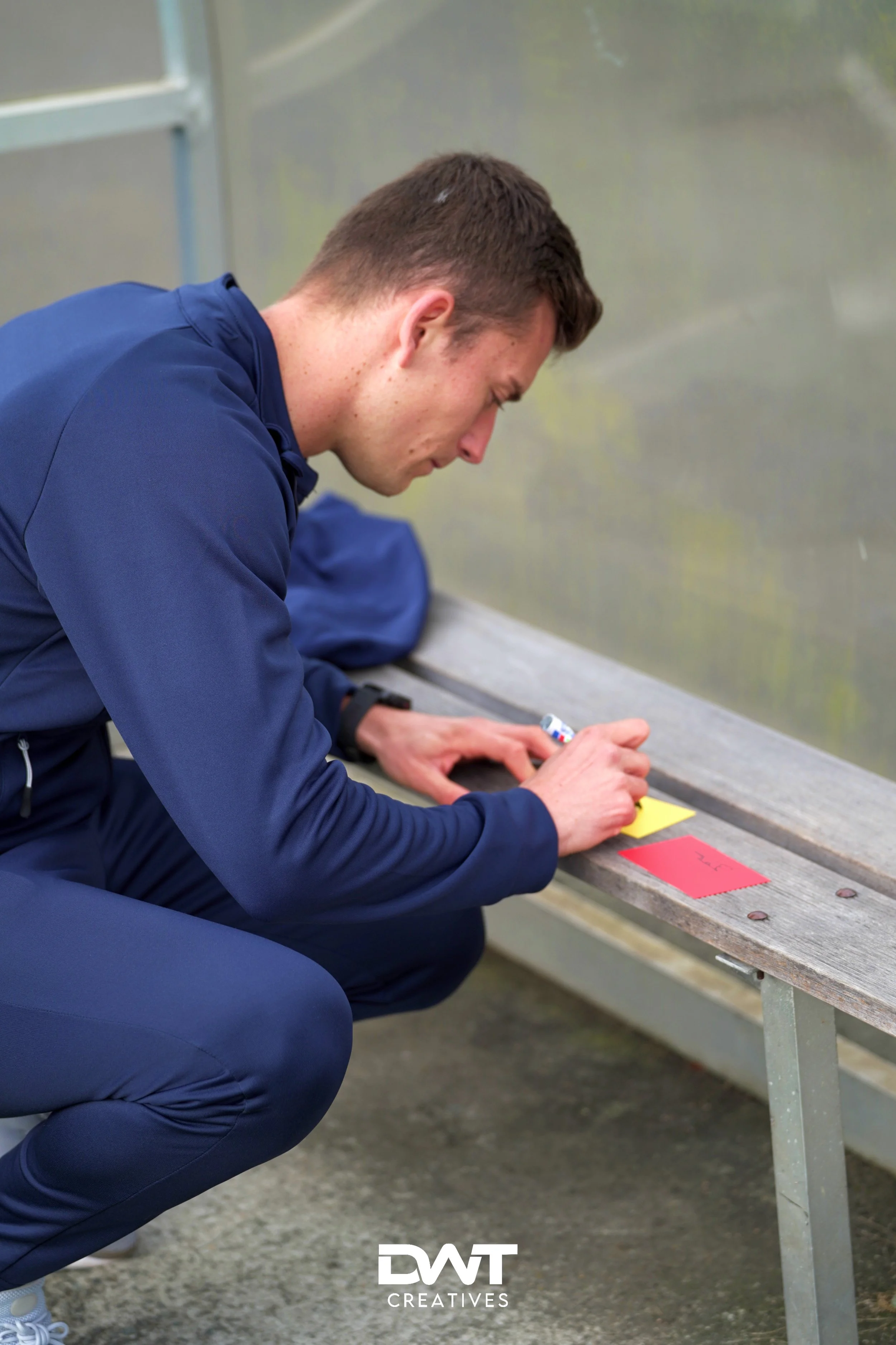 Een persoon in een donkerblauw sportpak die op een bankje zit en iets schrijft op een gekleurd papier.
