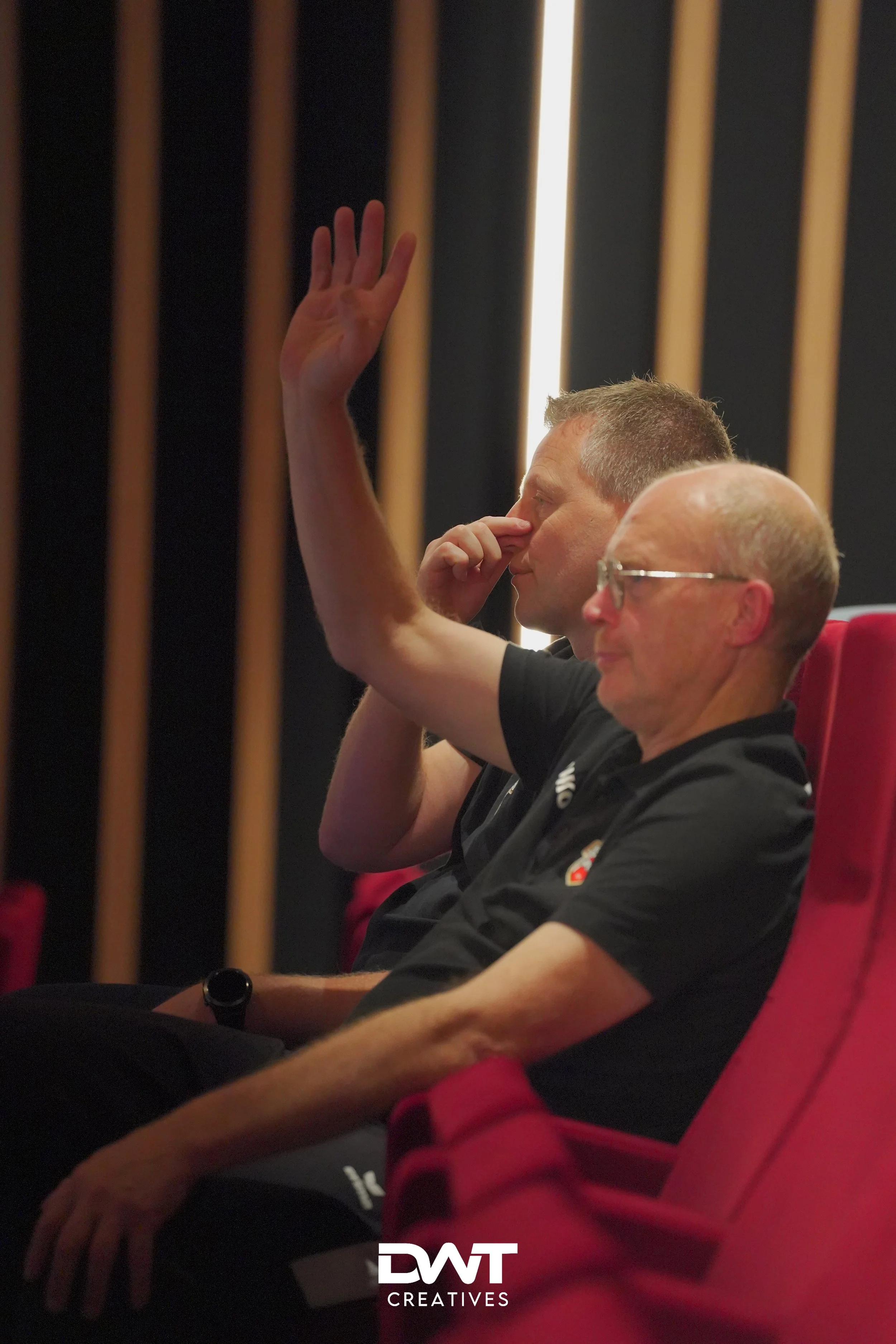 Twee mannen zitten in rode theaterstoelen; één steekt zijn hand op terwijl de andere zijn gezicht aanraakt.