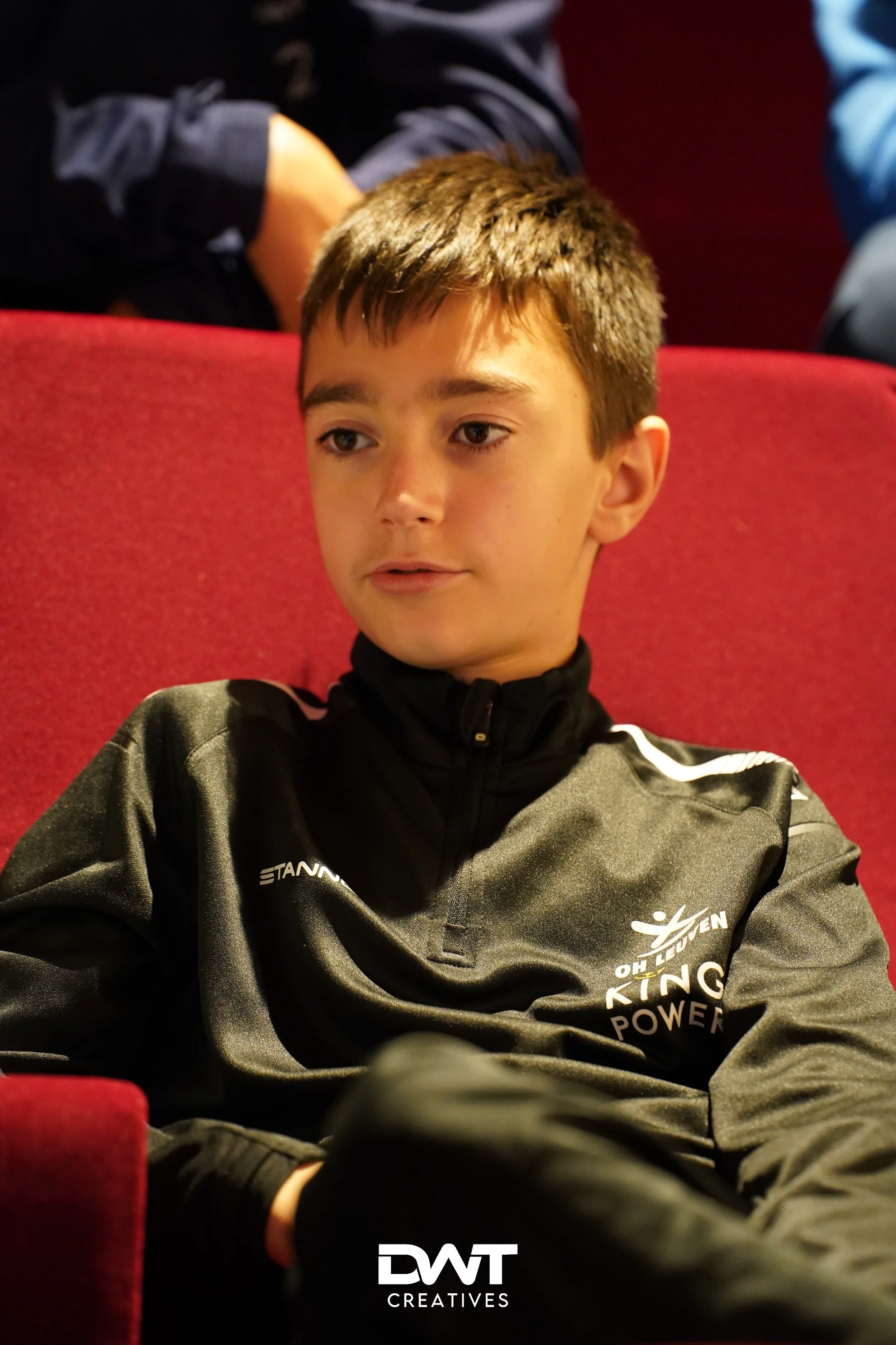 Jongen in zwart sportjack zittend op rode stoel; logo "OHL KEUVEN KING POWER" zichtbaar. "DWT CREATIVES" tekst onderaan.