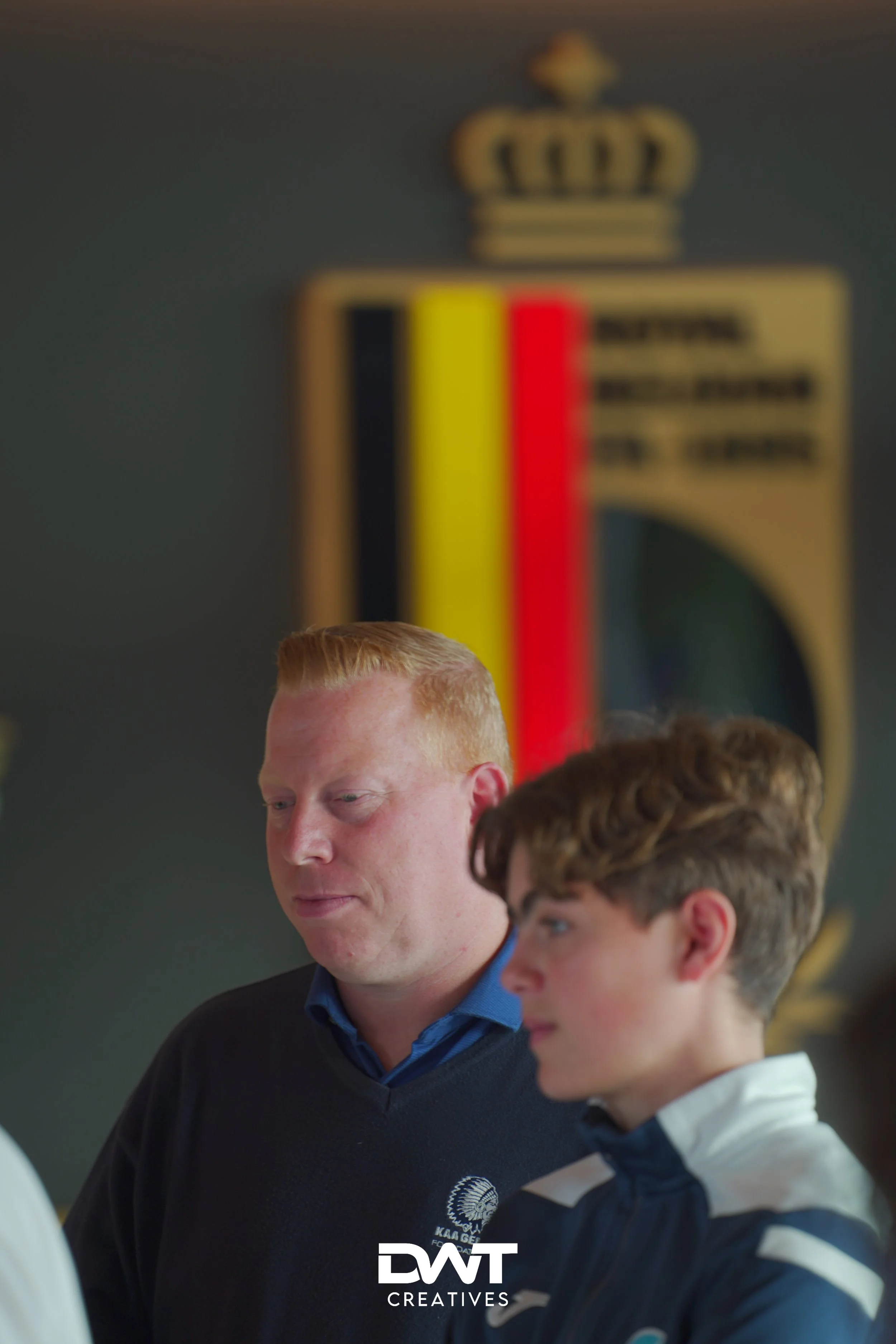 Twee personen staan voor een wazige achtergrond met een vlag en een kroon. De focus ligt op een man links en een jongere persoon rechts, beide in sportieve kleding. Onderaan de foto is het logo van DWT Creatives zichtbaar.