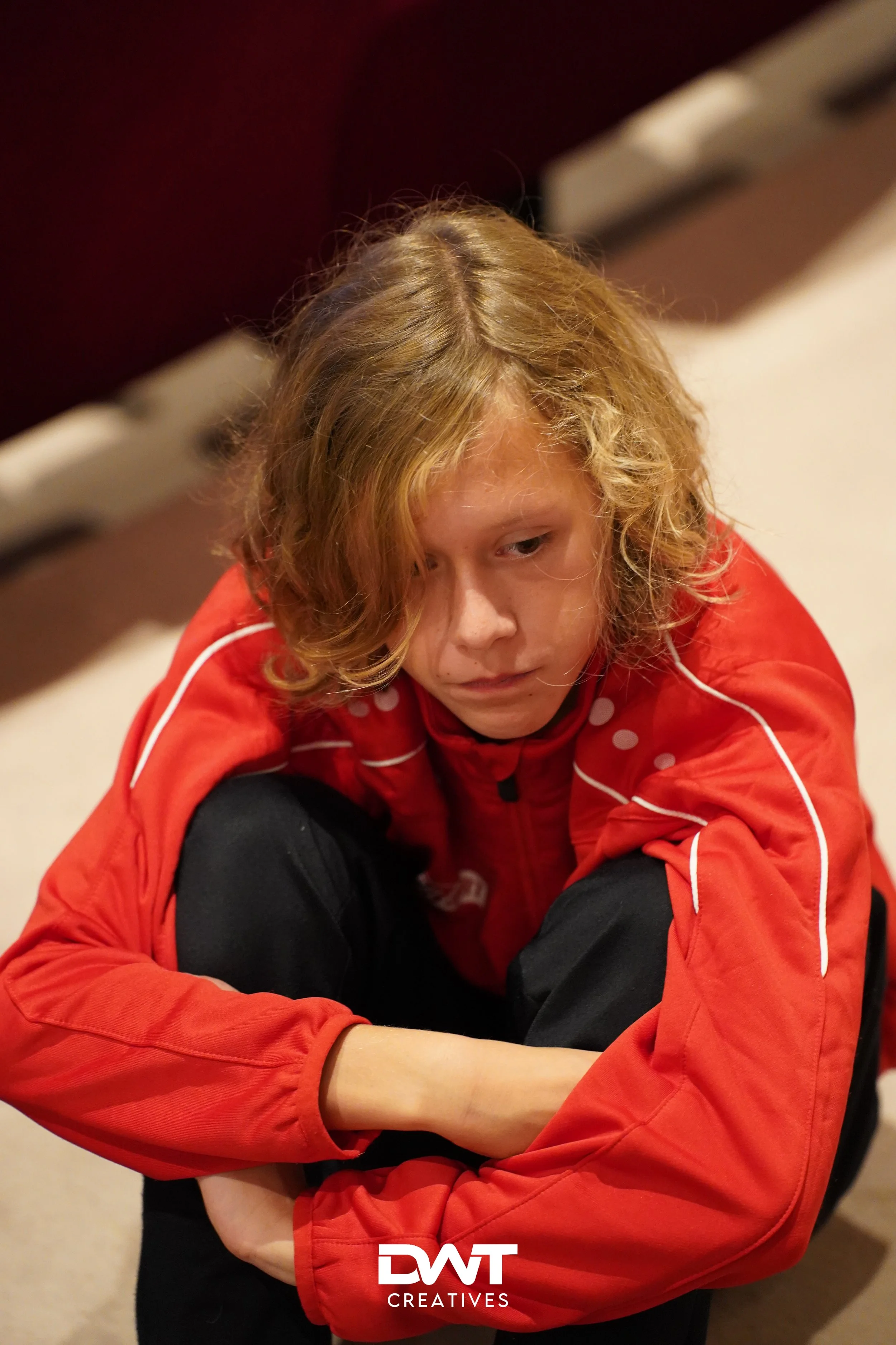 Een persoon met lang blond haar draagt een rode jas en zit op de grond met gebogen knieën.