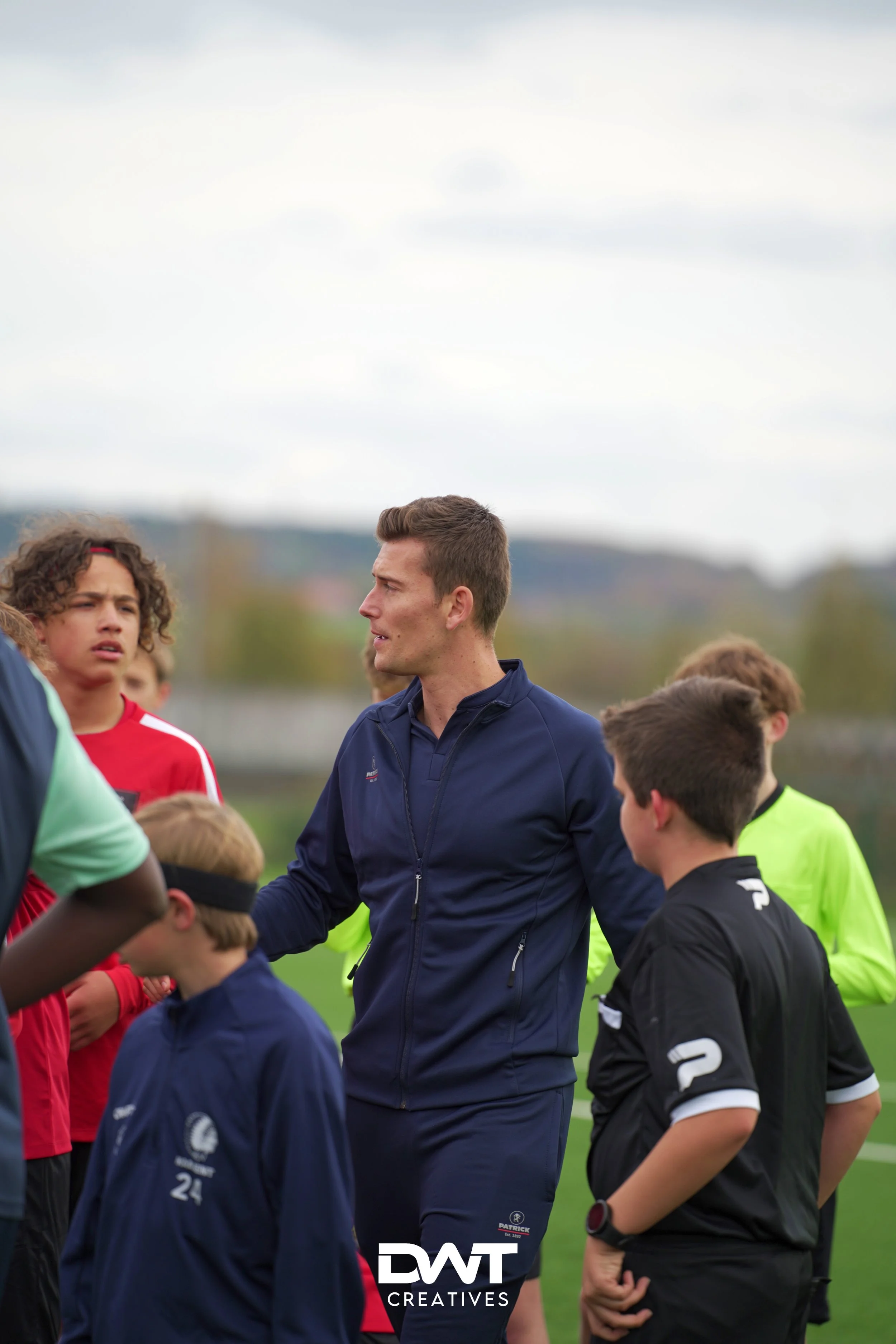 Voetbalcoach praat met jonge spelers op een sportveld. Het is bewolkt en spelers dragen sportkleding van verschillende kleuren.