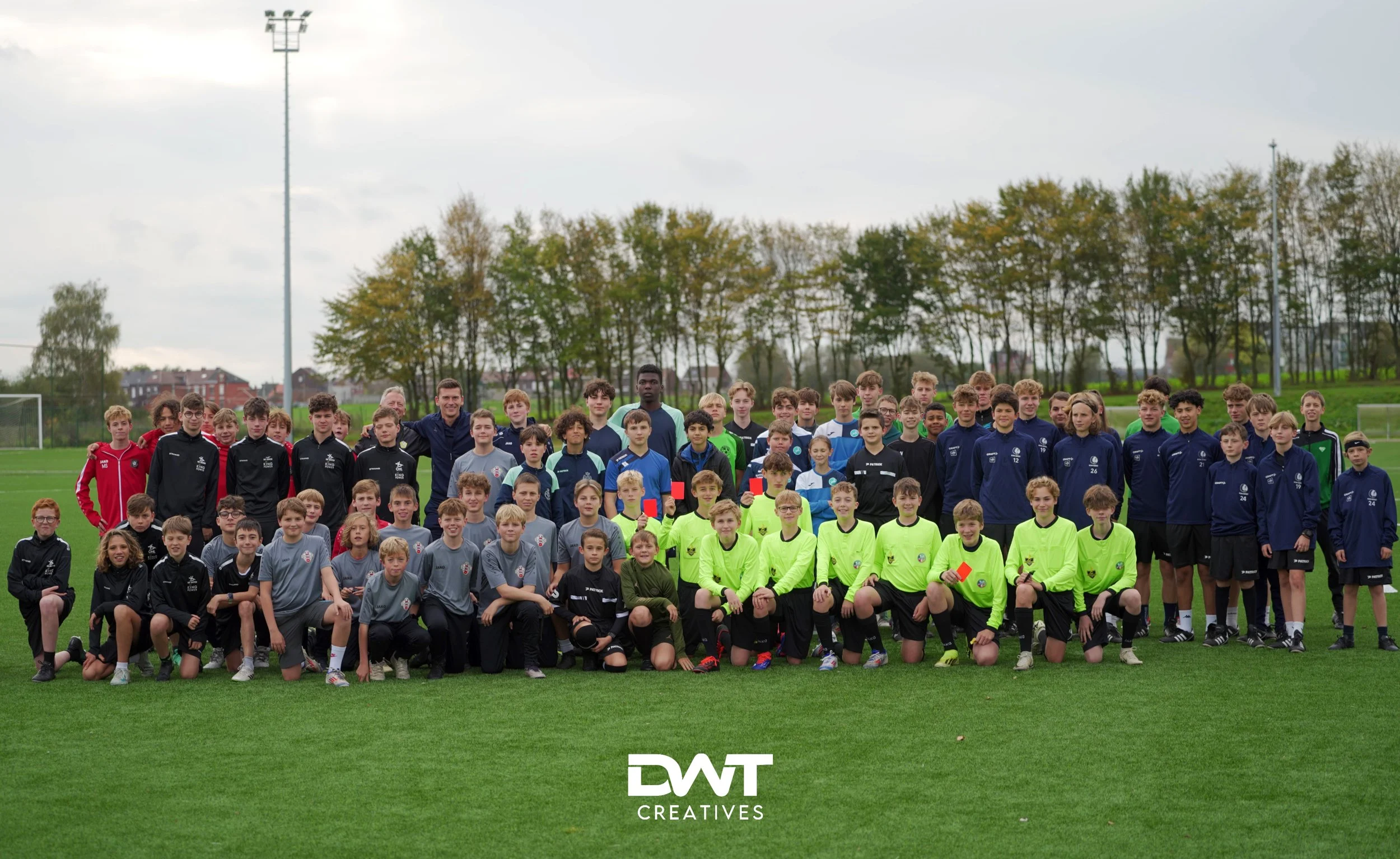 Groep jonge voetballers poseert op een sportveld met trainingspakken aan, omringd door bomen.