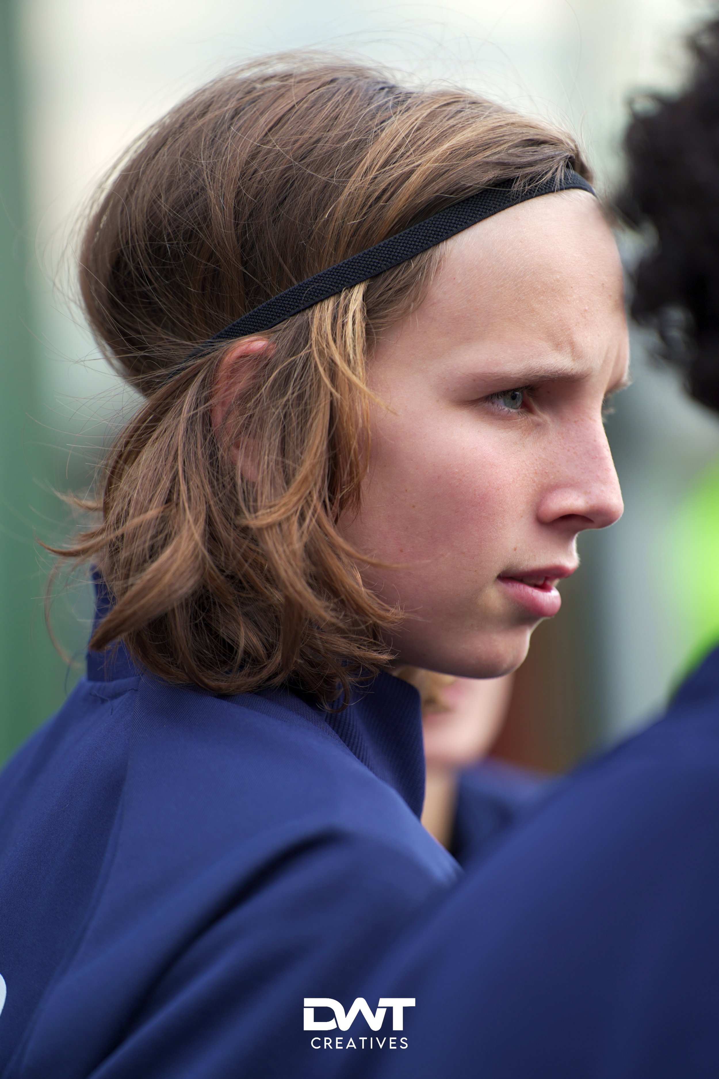 Jonge persoon met bruin haar en een hoofdband, gekleed in een blauwe outfit, kijkt geconcentreerd naar iets.