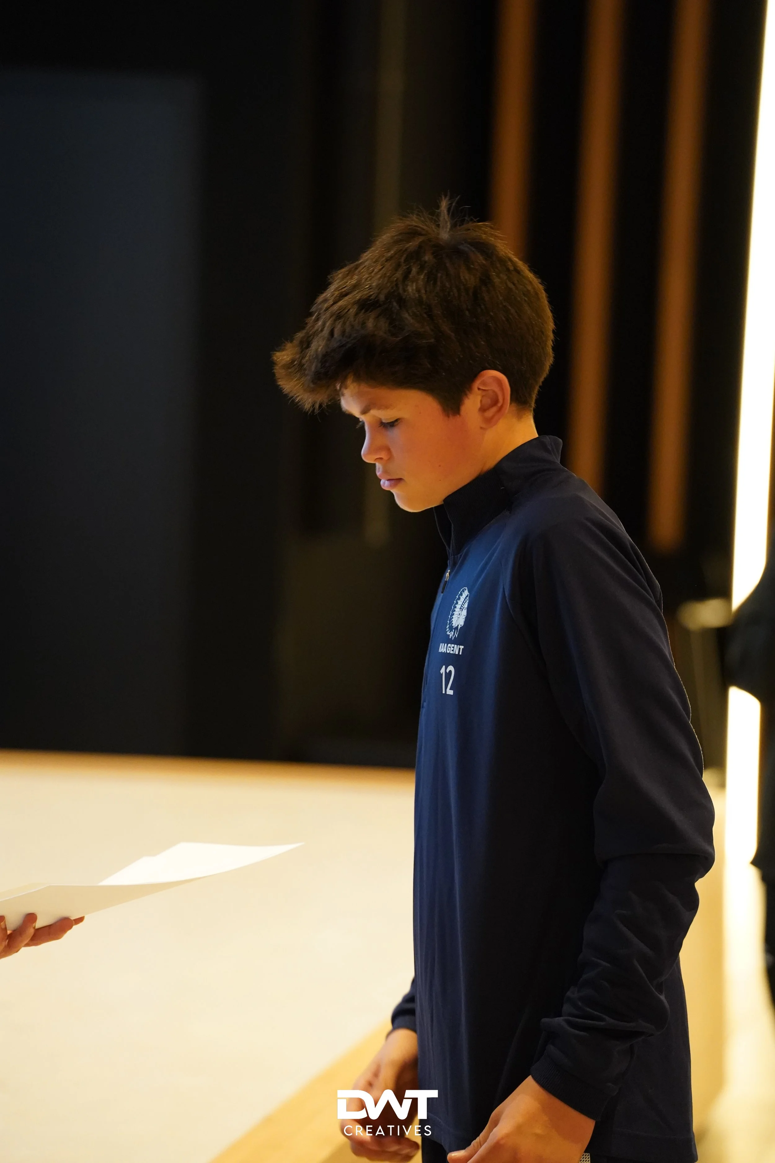 Jongen in blauwe trui kijkt naar papier dat door een ander persoon wordt vastgehouden in een binnenruimte. Op de trui staat "12" geschreven en het logo van DWT Creatives is zichtbaar.