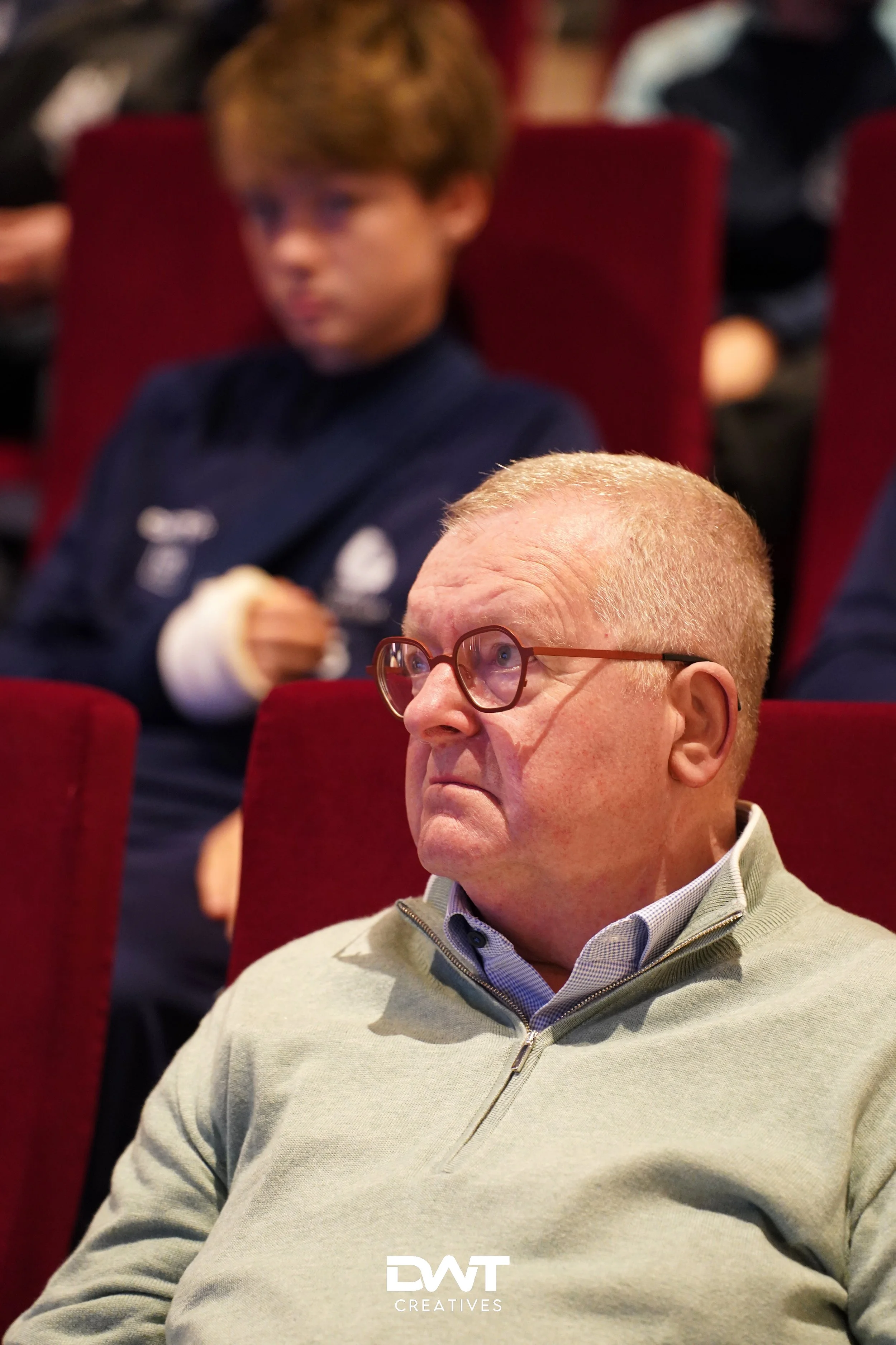 Twee mannen in een publiek, de voorste draagt een bril en een lichtgroene trui. Achter hen is een jonge man in een donkerblauw shirt te zien. Ze zitten in rode theaterstoelen.