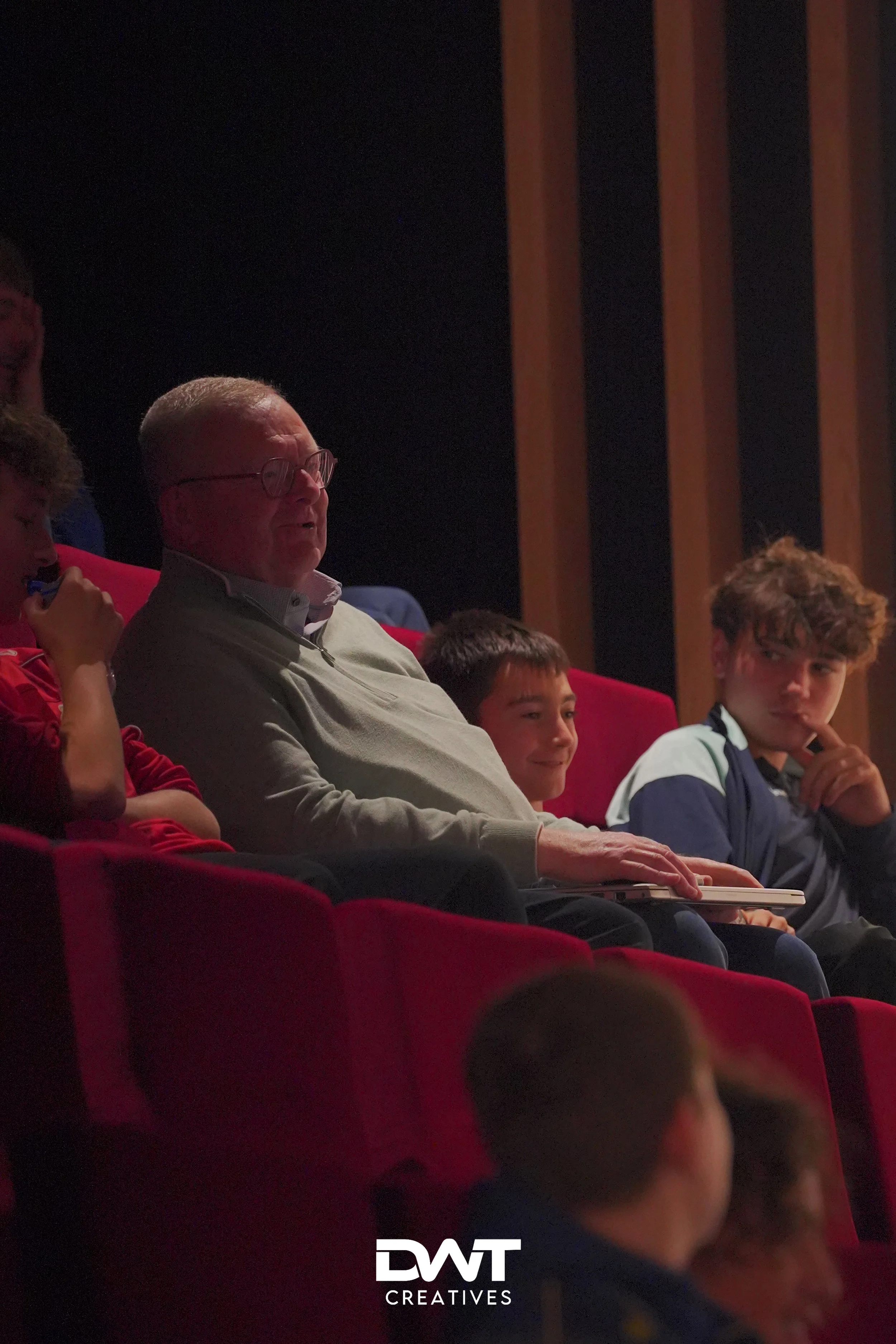 Een groep mensen zit samen in een theater of auditorium, op rode stoelen. Een oudere man met bril zit in het midden, omringd door jongere mensen. De sfeer lijkt ontspannen en de achtergrond is donker.