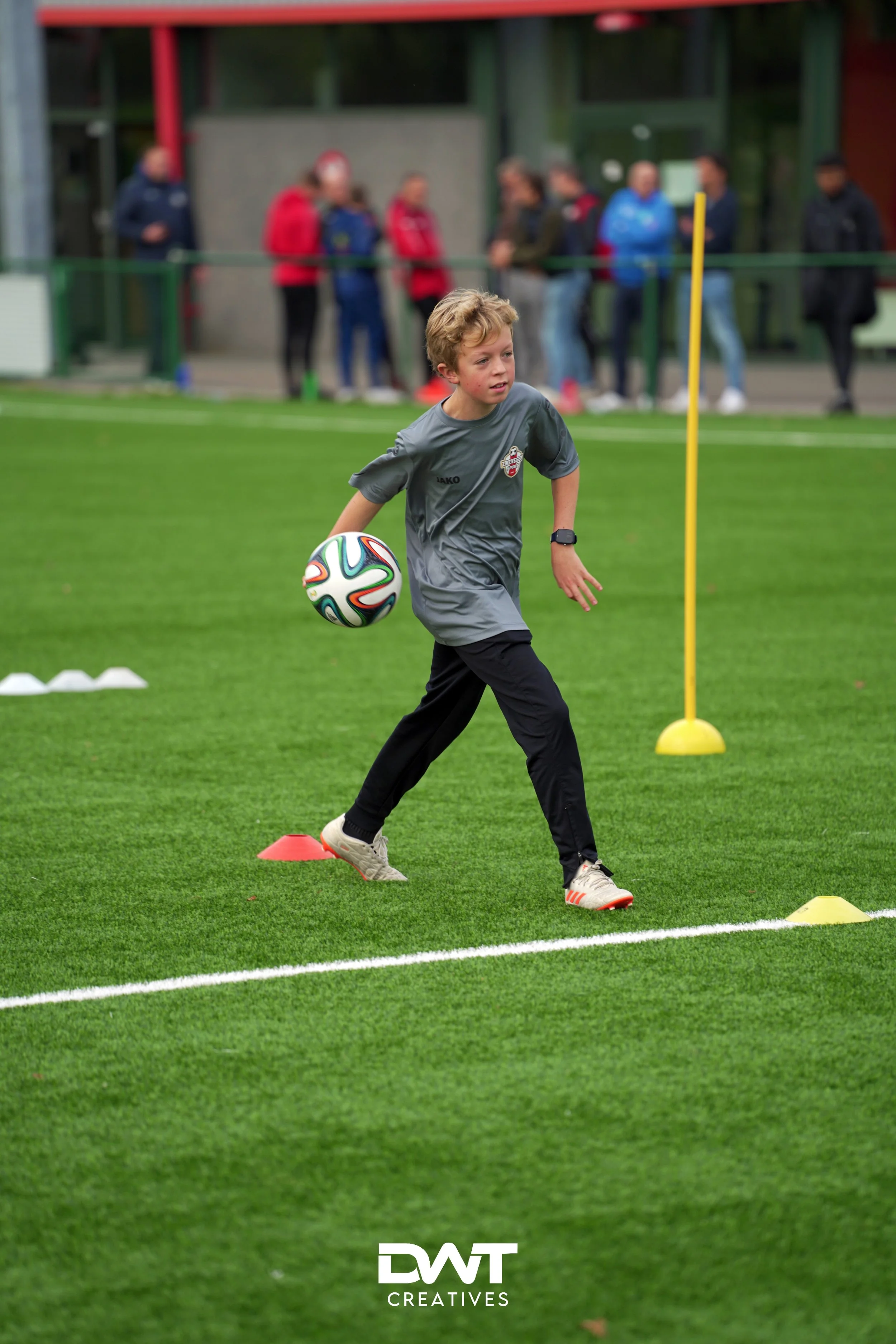 Kind voetbalt op veld met kegels, bal vast. Andere mensen op achtergrond bij voetbaltraining.