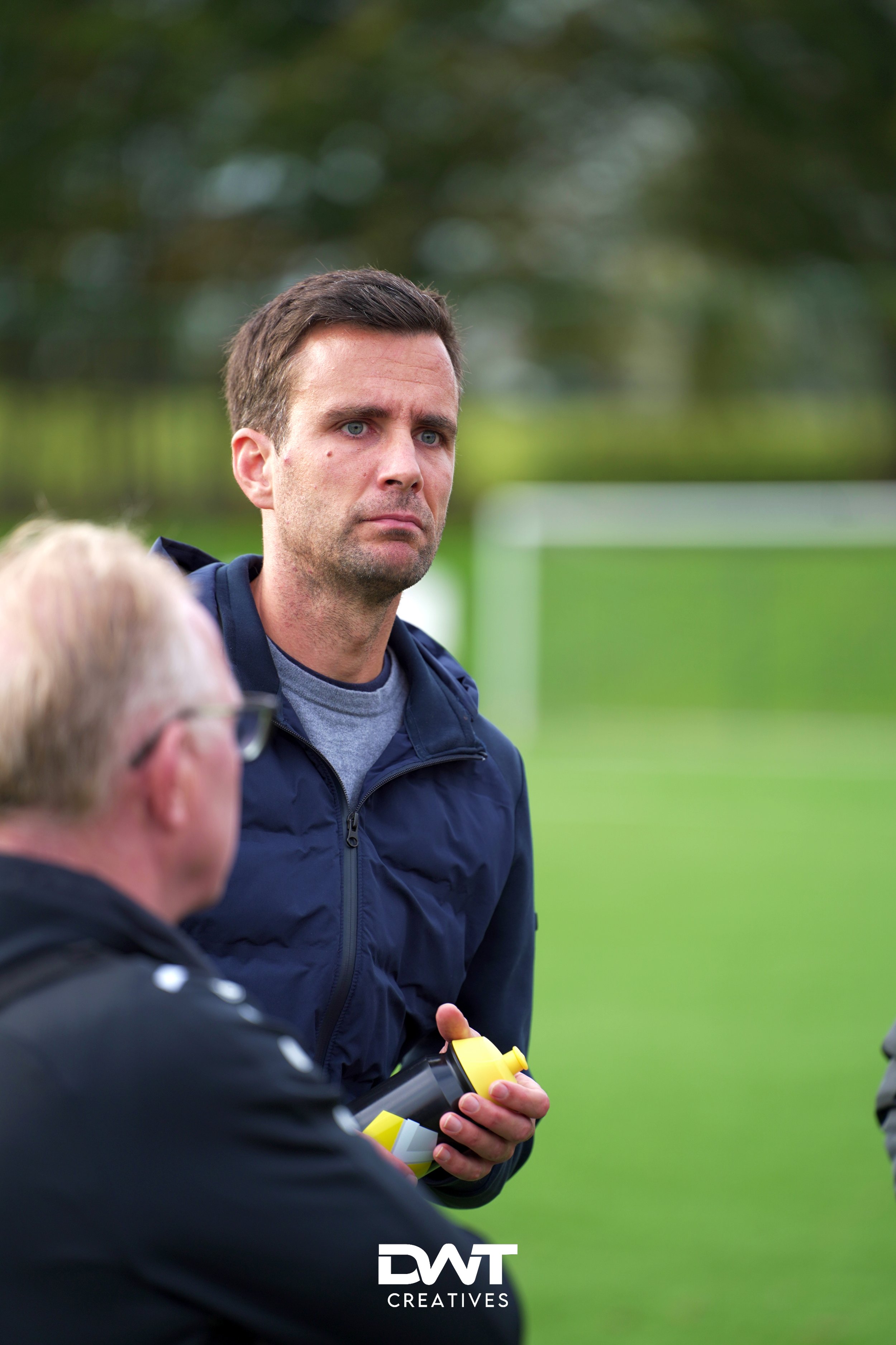 Twee mannen op een sportveld, één met een donkerblauwe jas en een bidon in zijn hand. Achtergrond is vaag met groen gras en een doel, suggererend een voetbalveld. Logo 'DWT Creatives' onderaan.