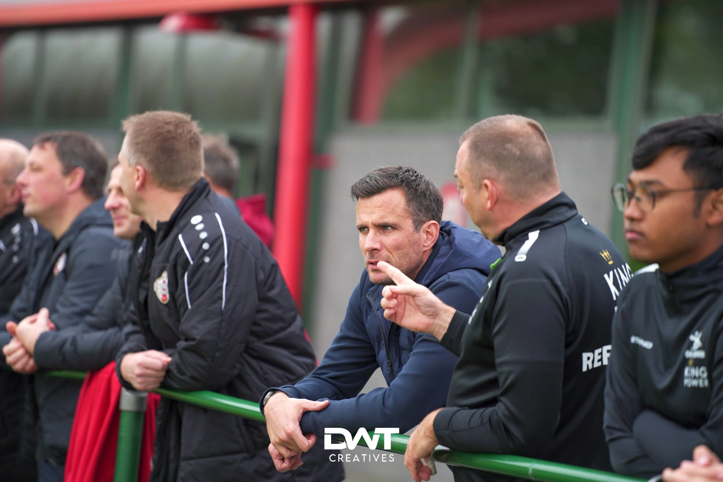 Groep mannen die langs een groene reling kijken, sommigen met sportkleding. Een man gebaart met zijn hand. Op de achtergrond een gebouw en groenblauwe tinten.