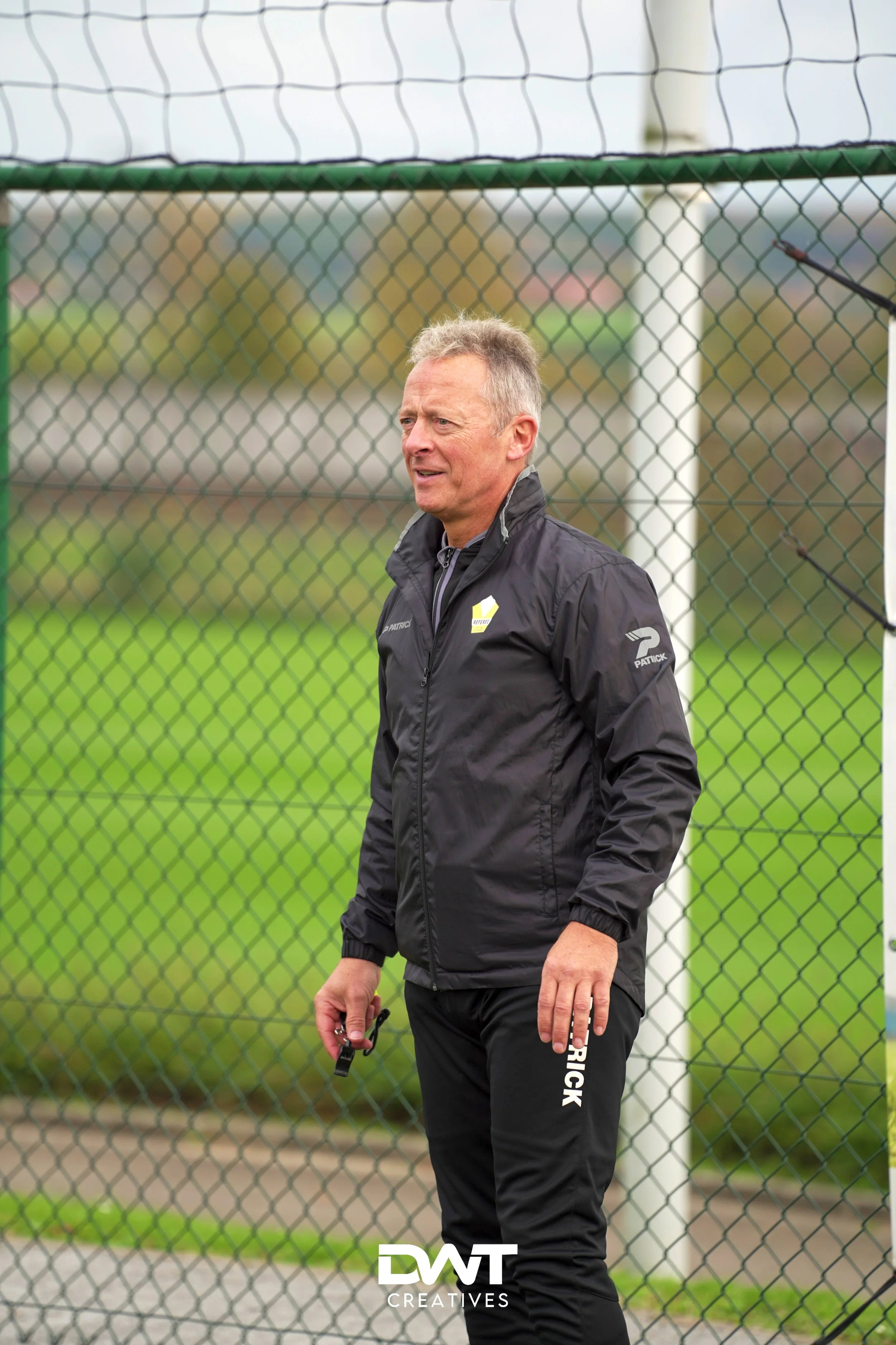 Man in zwart trainingspak met fluitje bij hekwerk op sportveld.