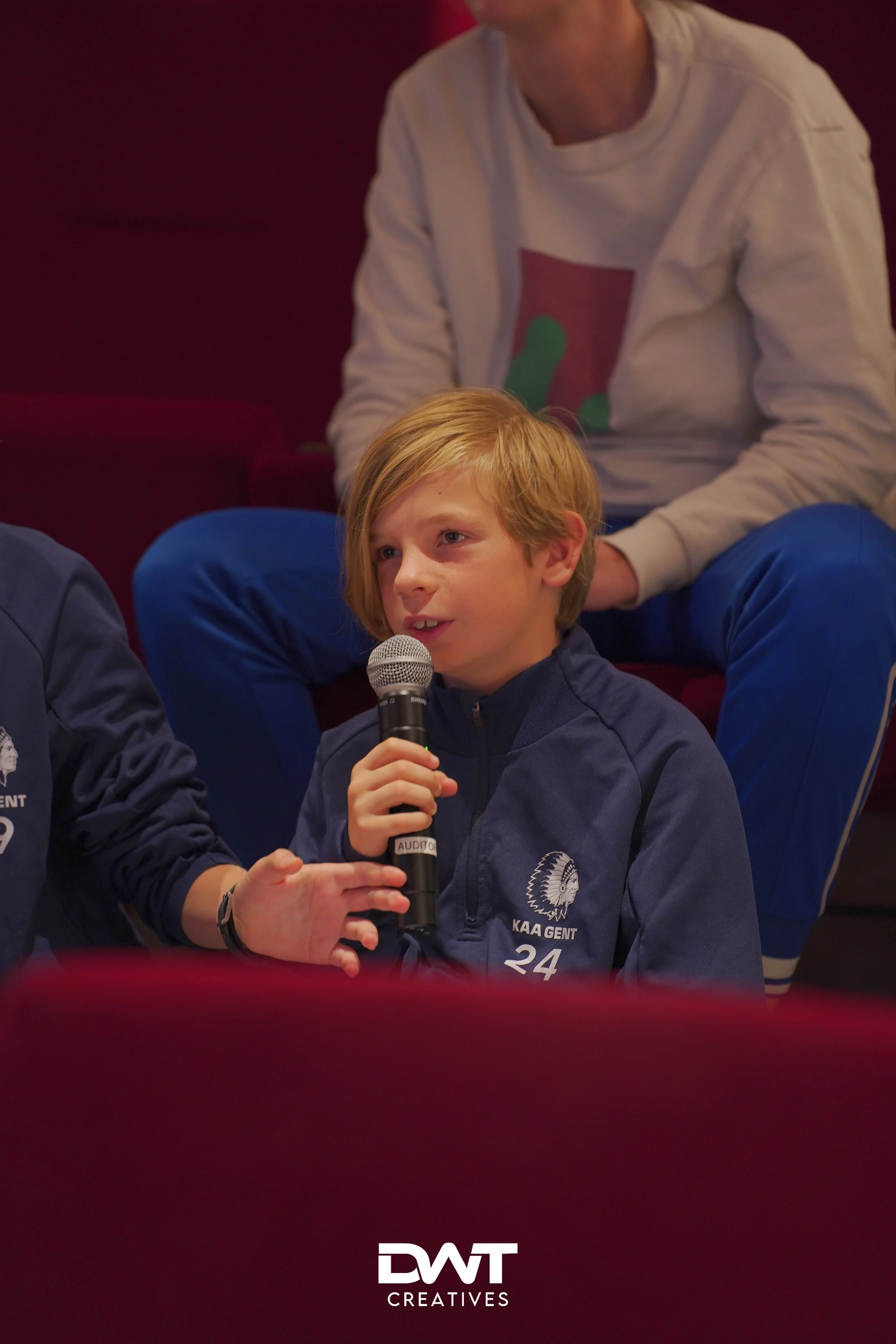 Een jongen spreekt in een microfoon, zittend op een rode bank. Hij draagt een blauw KAA Gent trainingsjack. Op de achtergrond zit een persoon, deels zichtbaar.