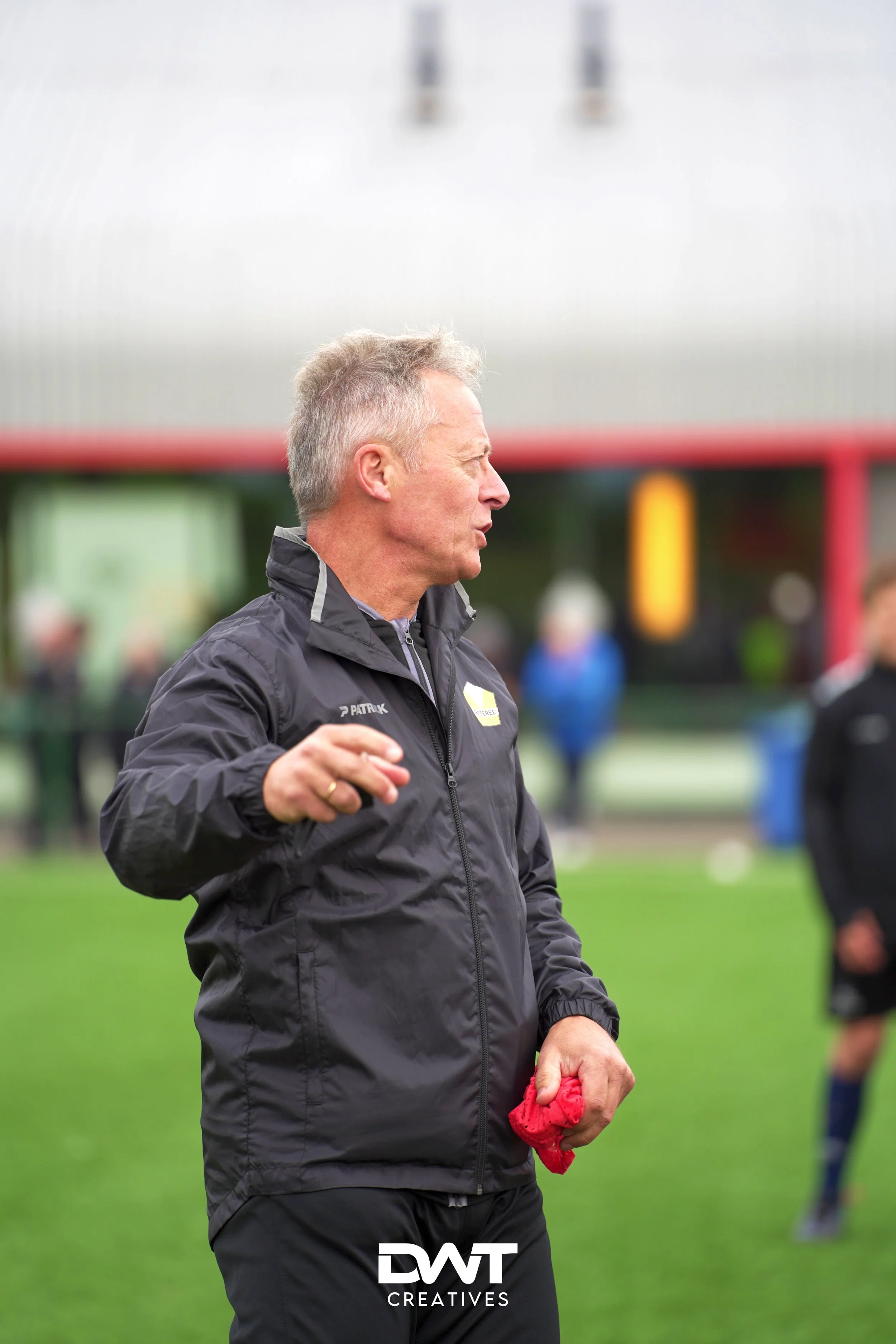 Man in zwarte jas op sportveld, wijzend met hand en rode doek vasthoudend.