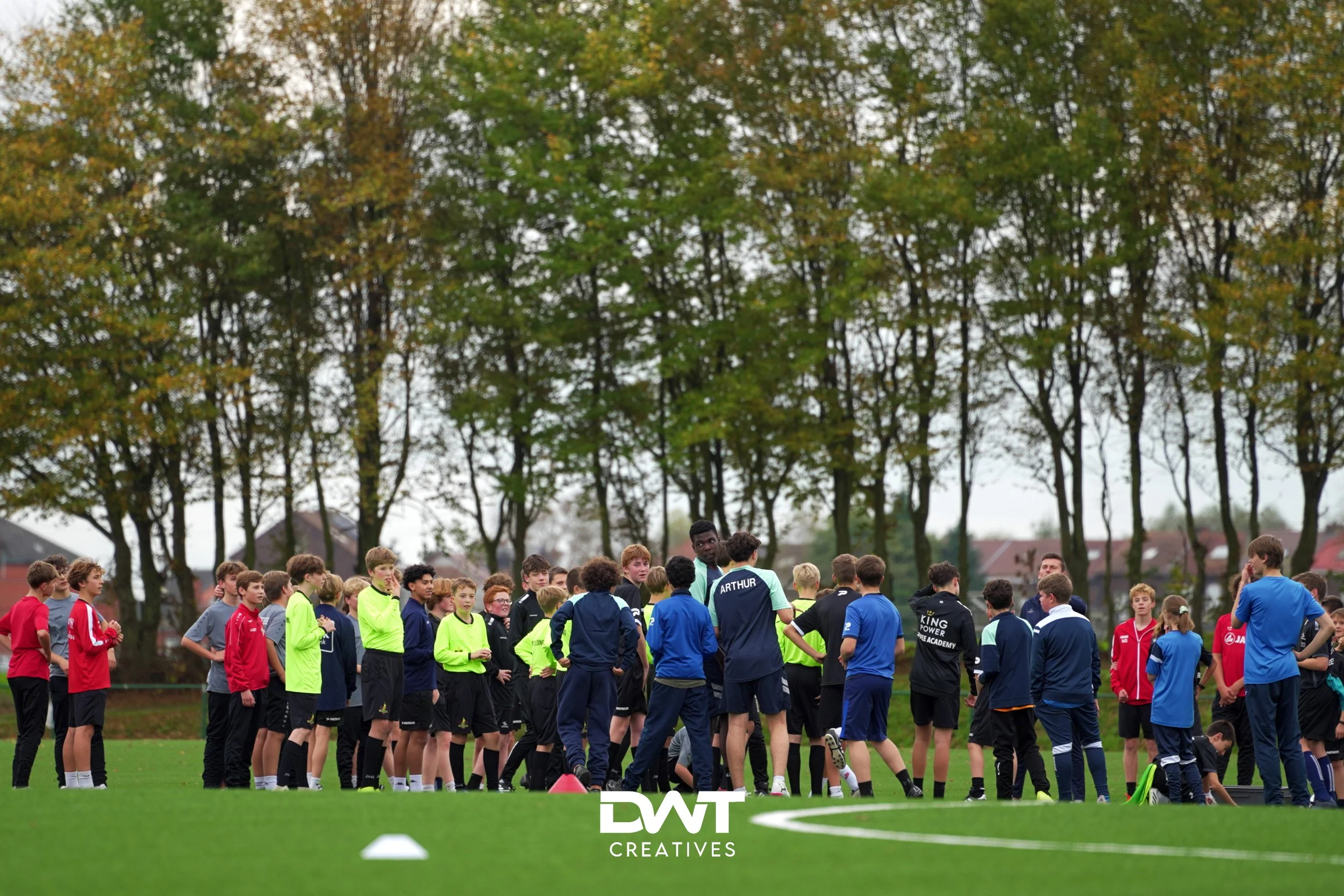 Een groep kinderen en jongeren staat op een grasveld in sportkleding tijdens een voetbaltraining. Op de achtergrond zijn bomen zichtbaar.