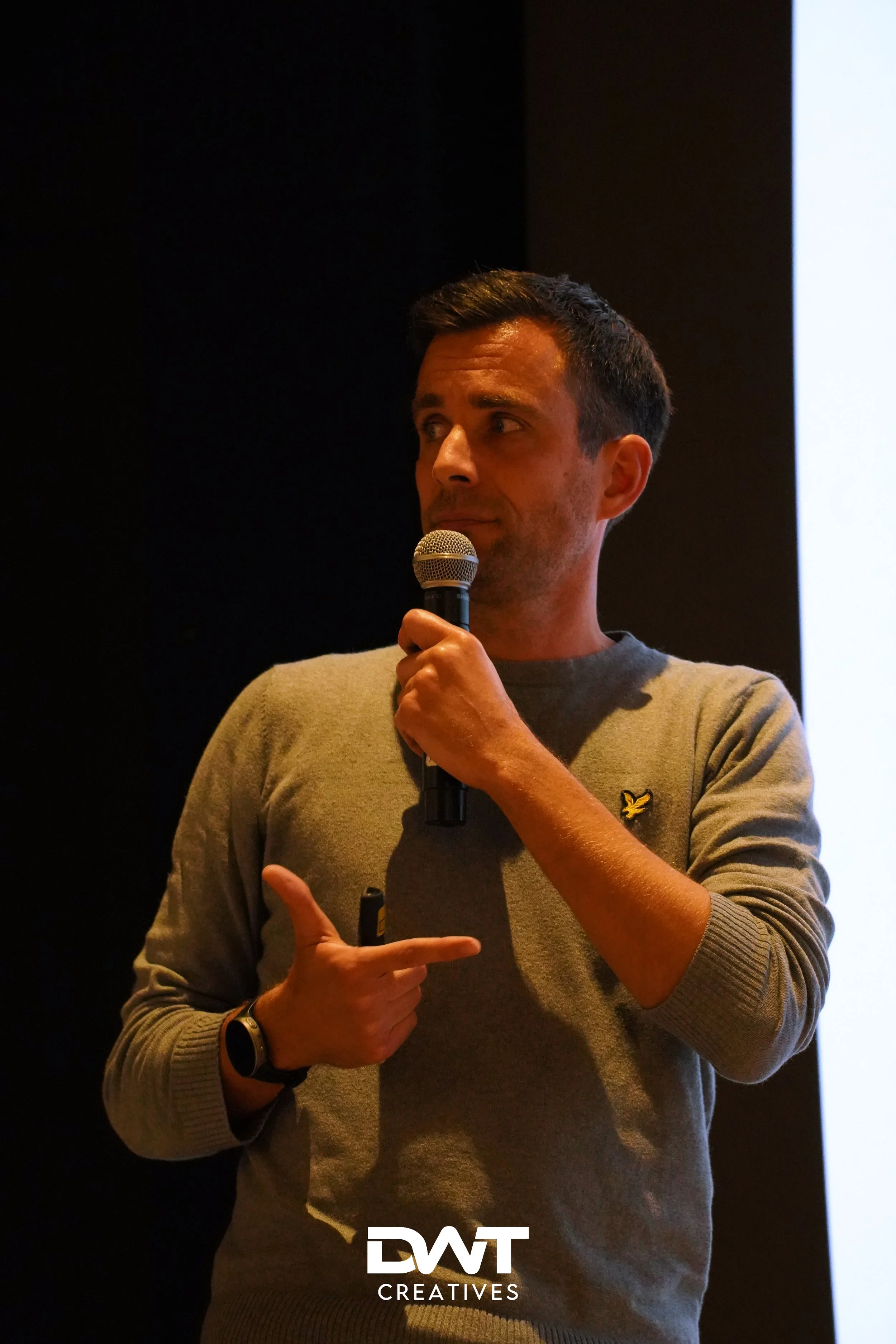 Man met microfoon tijdens een presentatie in een donkergrijs trui.