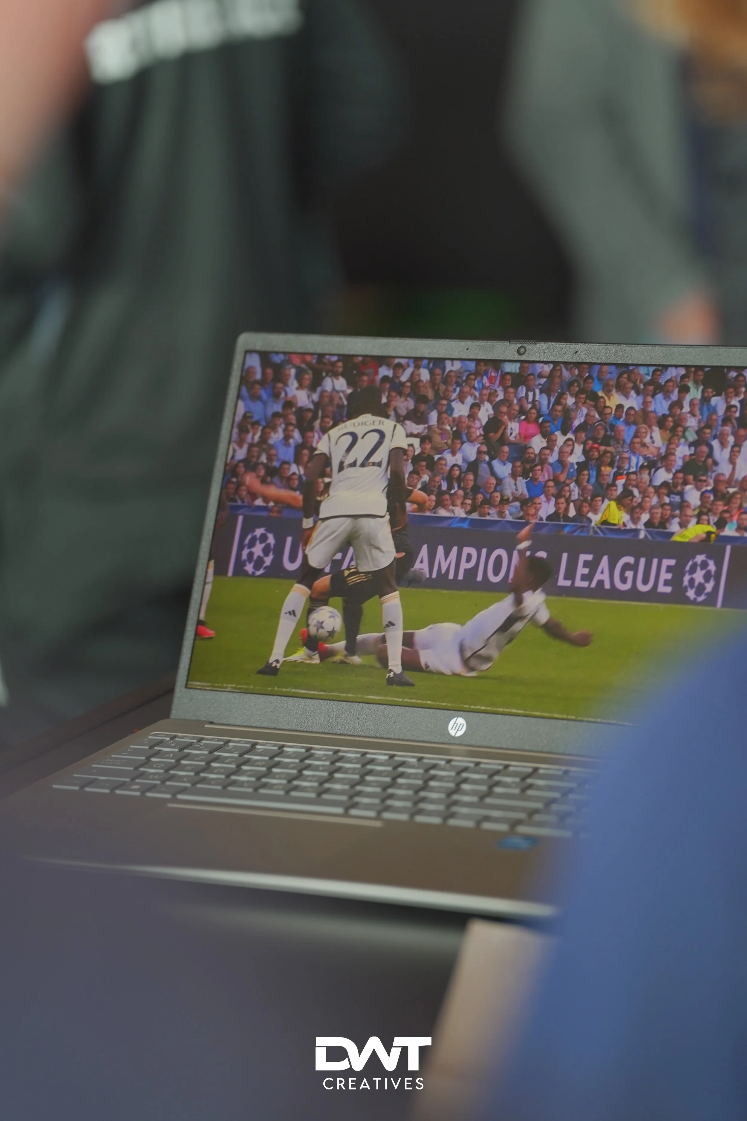 Close-up van een laptop waarop een voetbalwedstrijd van de UEFA Champions League wordt gestreamd, met spelers op het scherm en het publiek op de achtergrond. De laptop heeft een toetsenbord zichtbaar.