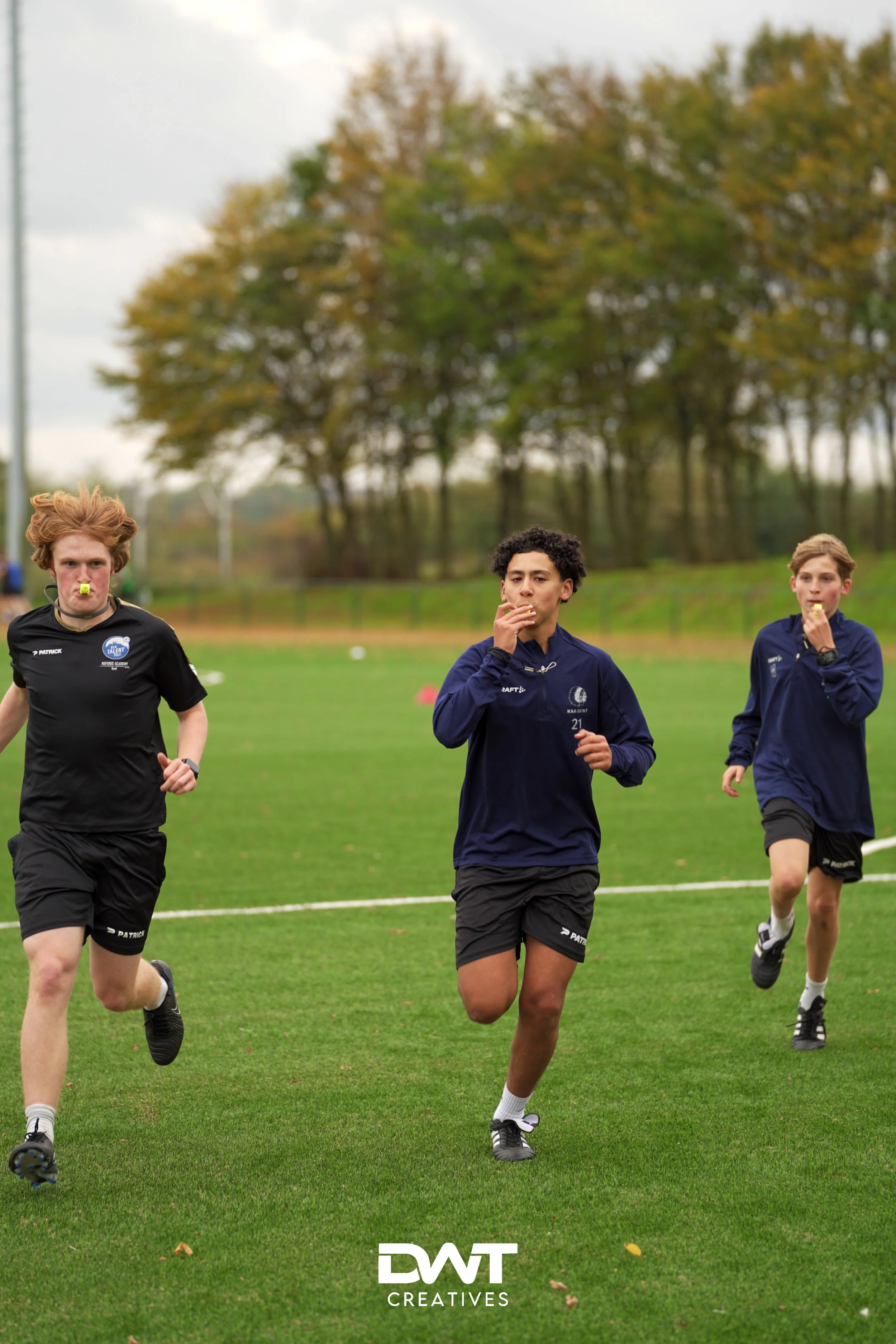 Drie jongens rennen op een sportveld met een groen grasveld. Ze dragen sportkleding: twee dragen donkerblauwe shirts, de ander een zwart shirt. Op de achtergrond zijn bomen zichtbaar.