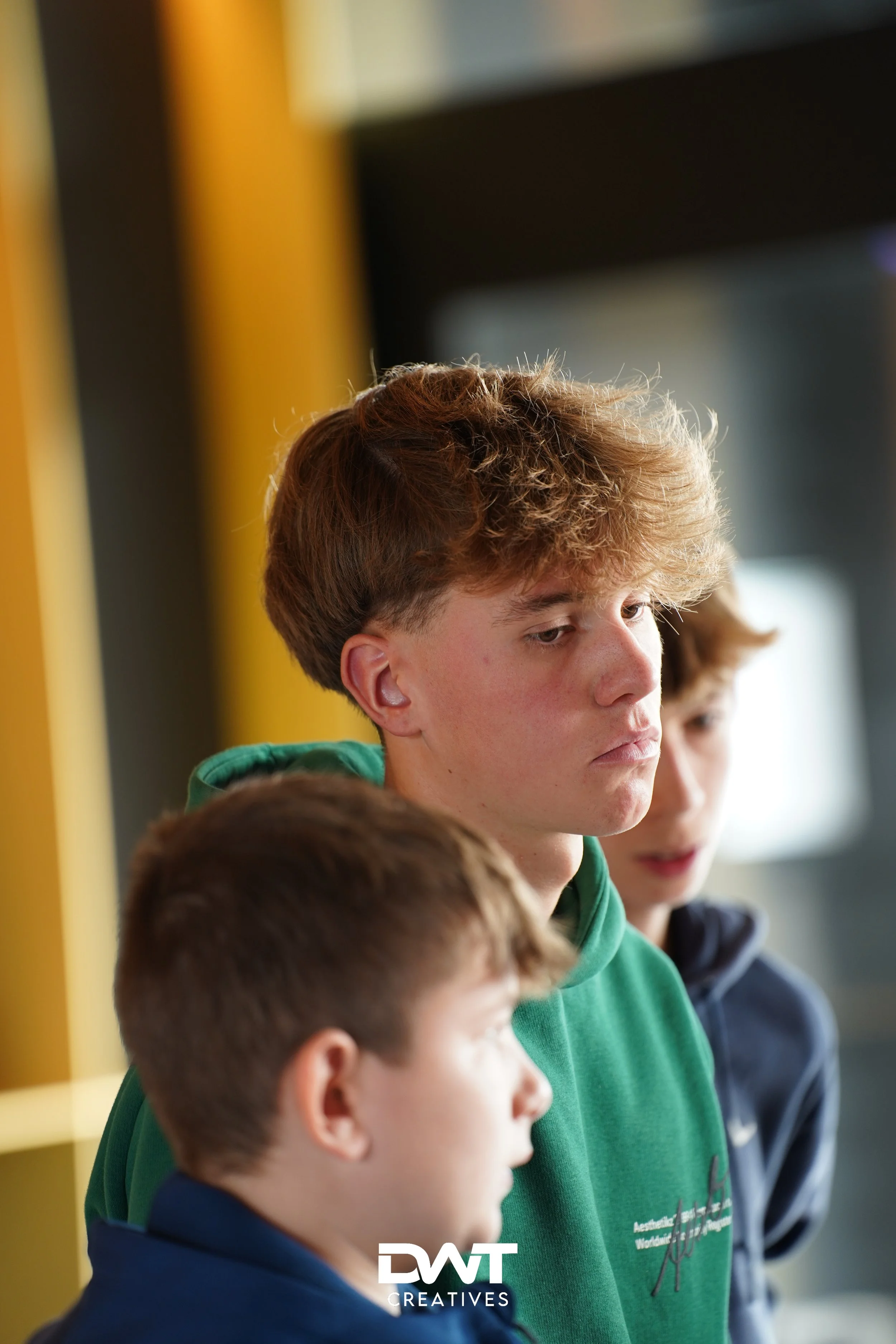 Drie jongeren, waarvan één een groene trui draagt, in gesprek met elkaar in een verlichte binnenruimte.