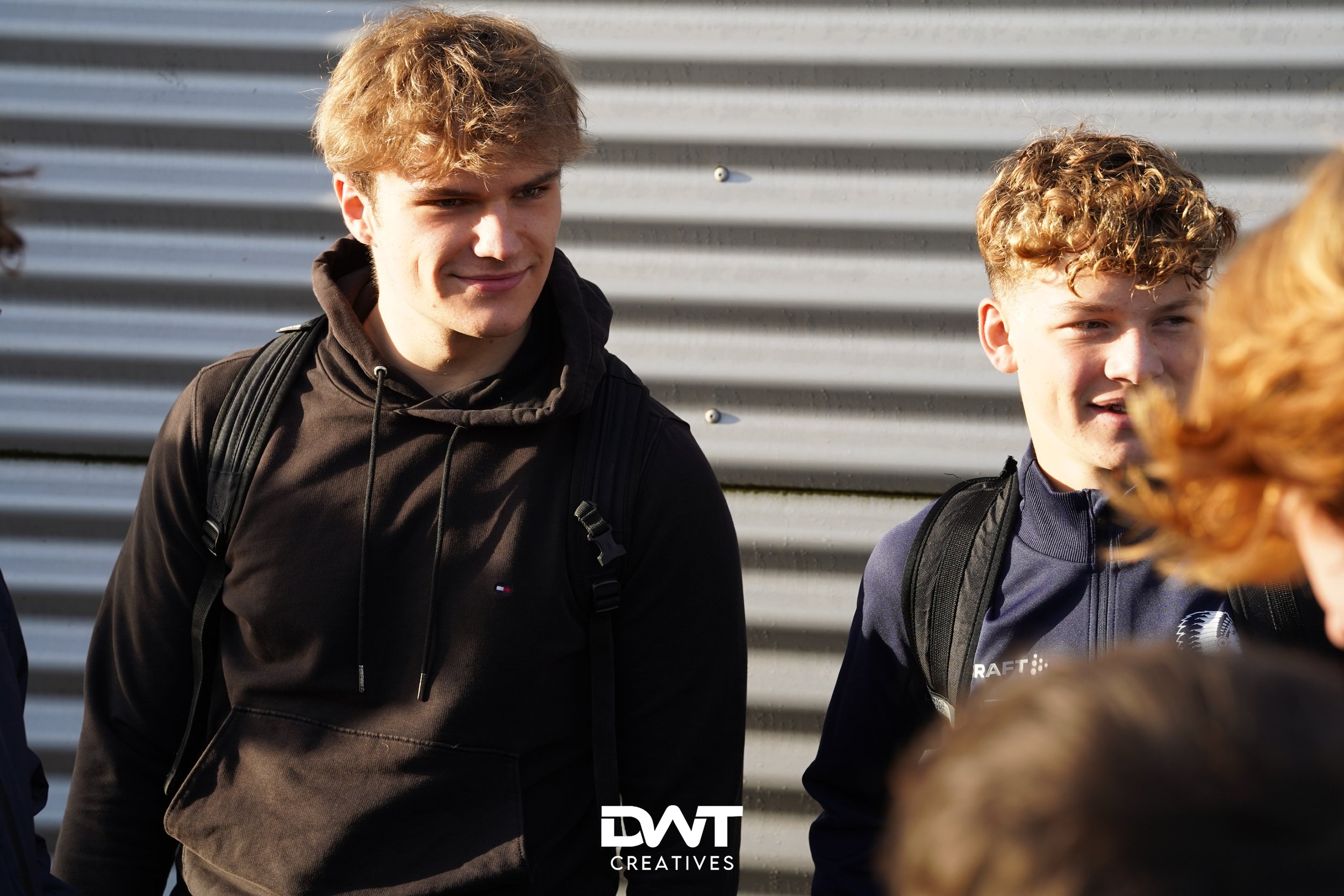 Twee jonge mannen met rugzakken staan buiten tegen een metalen achtergrond.