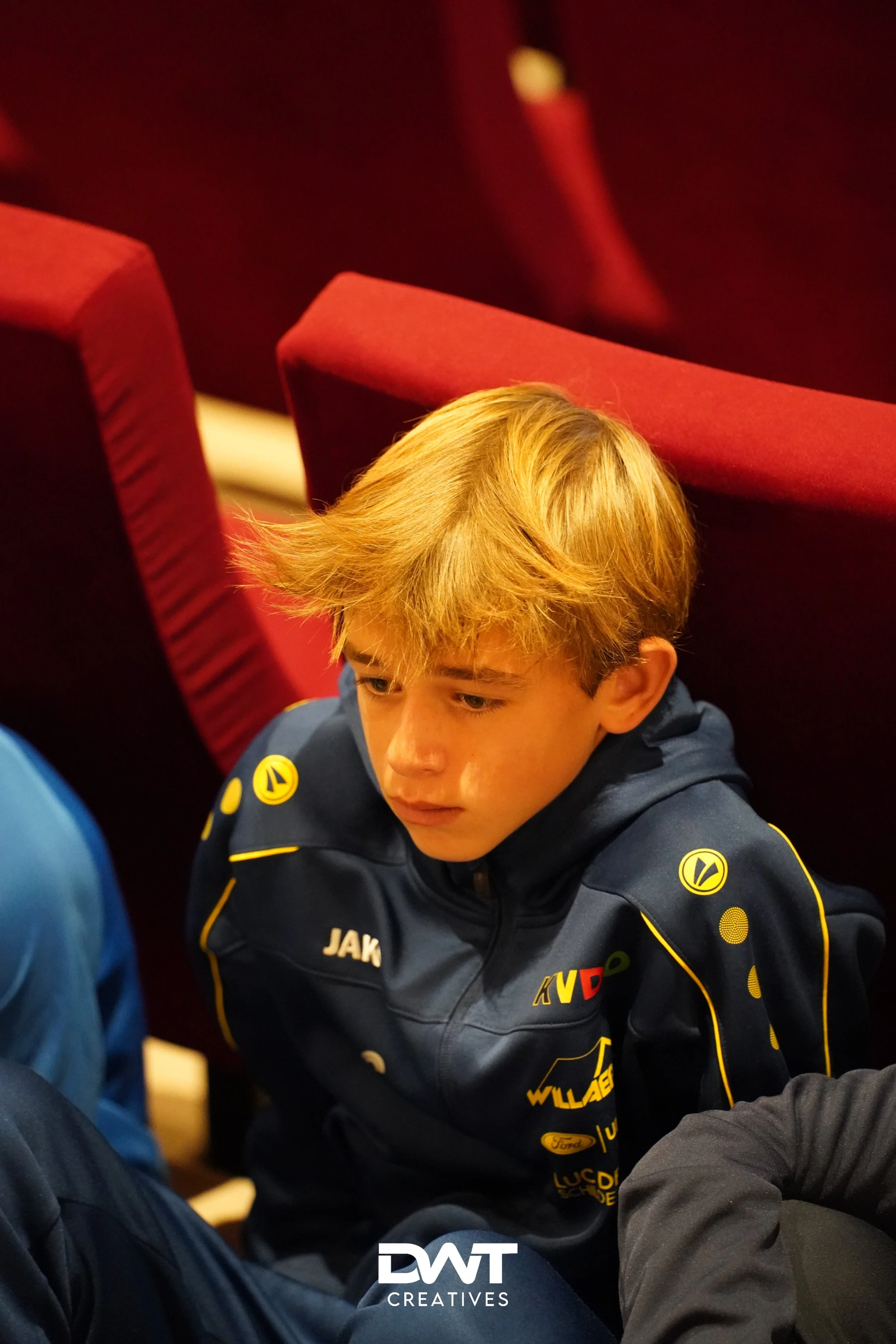 Jongen in blauwe sportkleding, zittend in een rode stoel, met geconcentreerde blik.