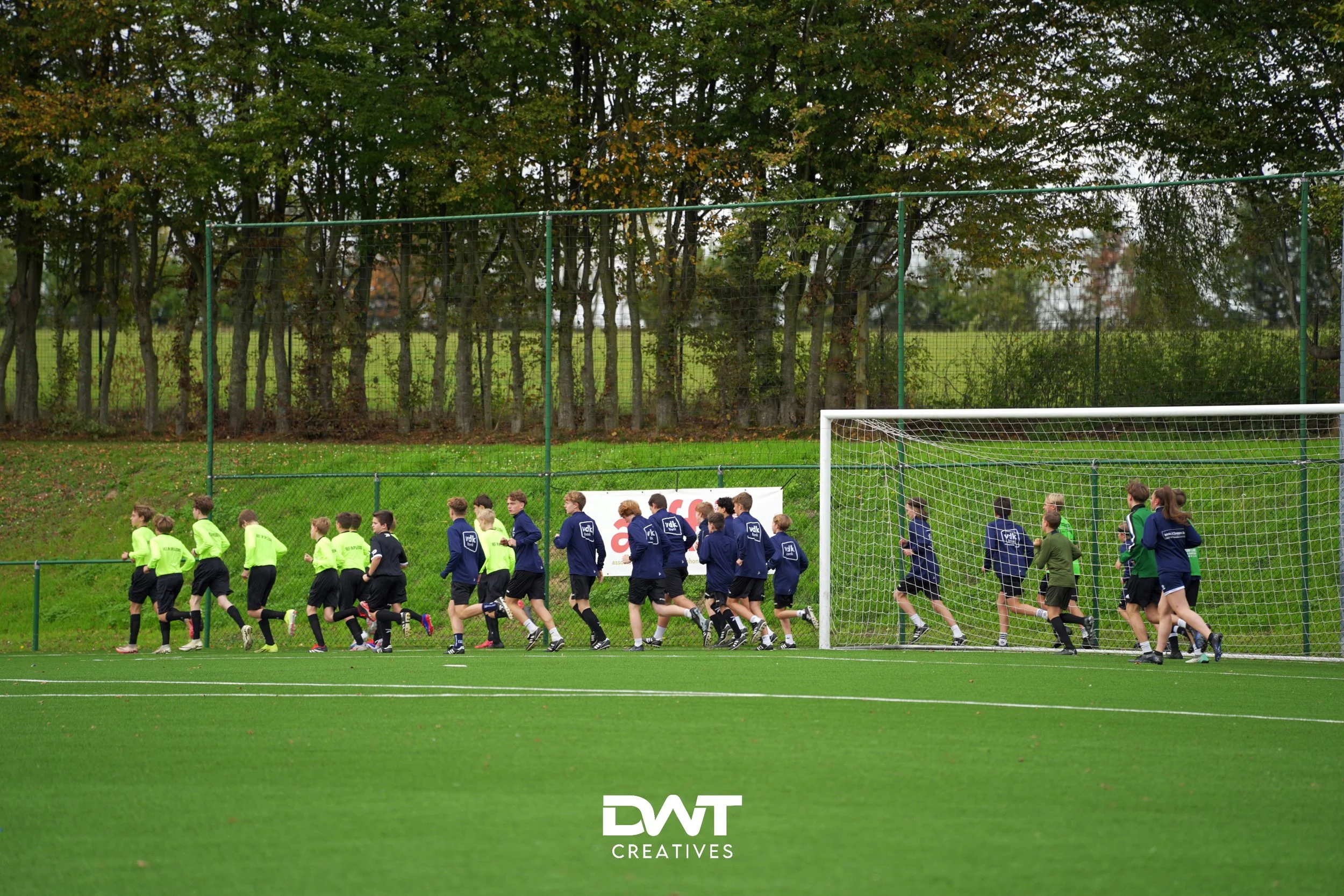 Groep kinderen rent over een voetbalveld in sportkleding, omringd door groen en bomen, met een doel op de achtergrond.