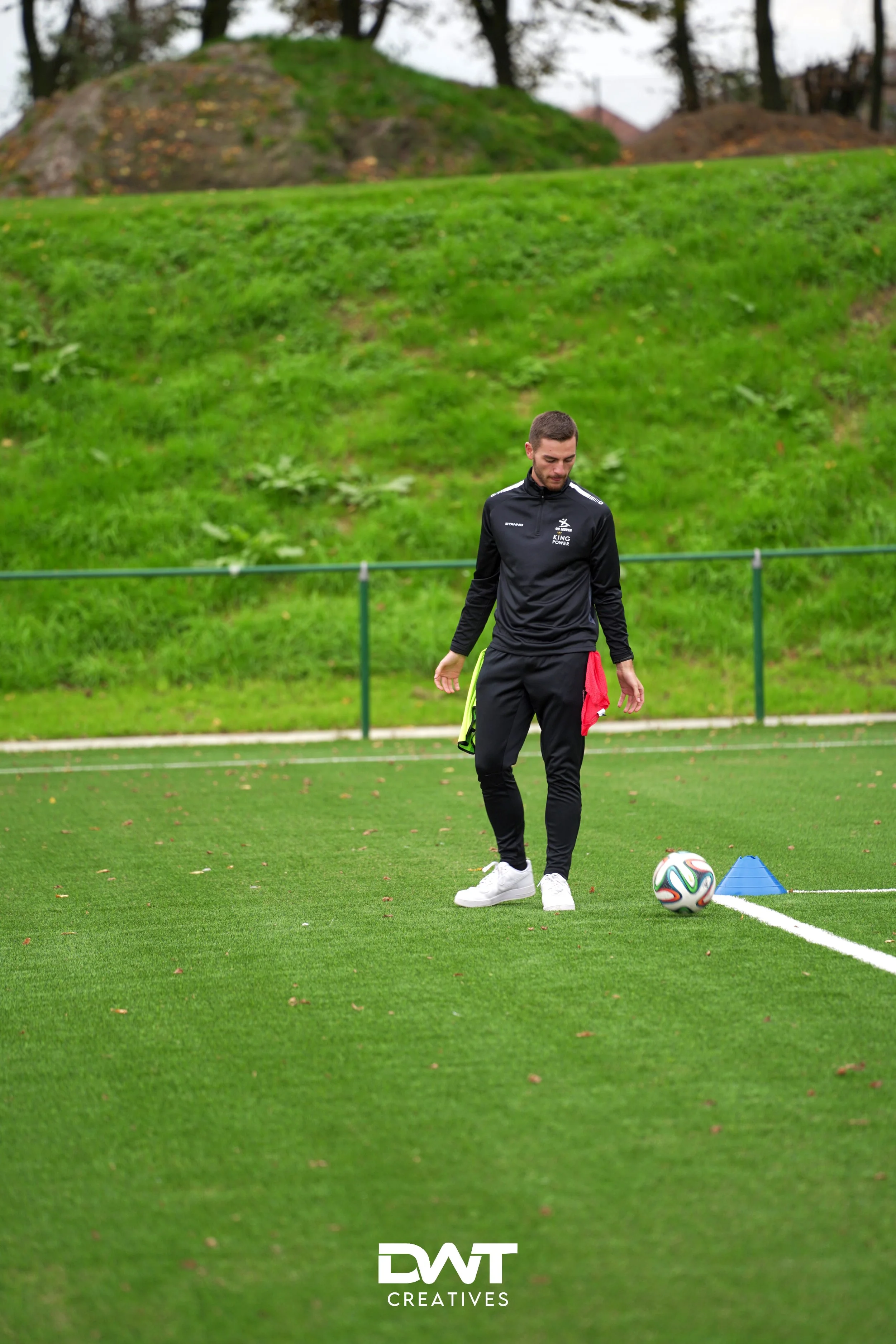 Man op voetbalveld met bal, in sportkleding, gras op achtergrond, trainingskegel zichtbaar.