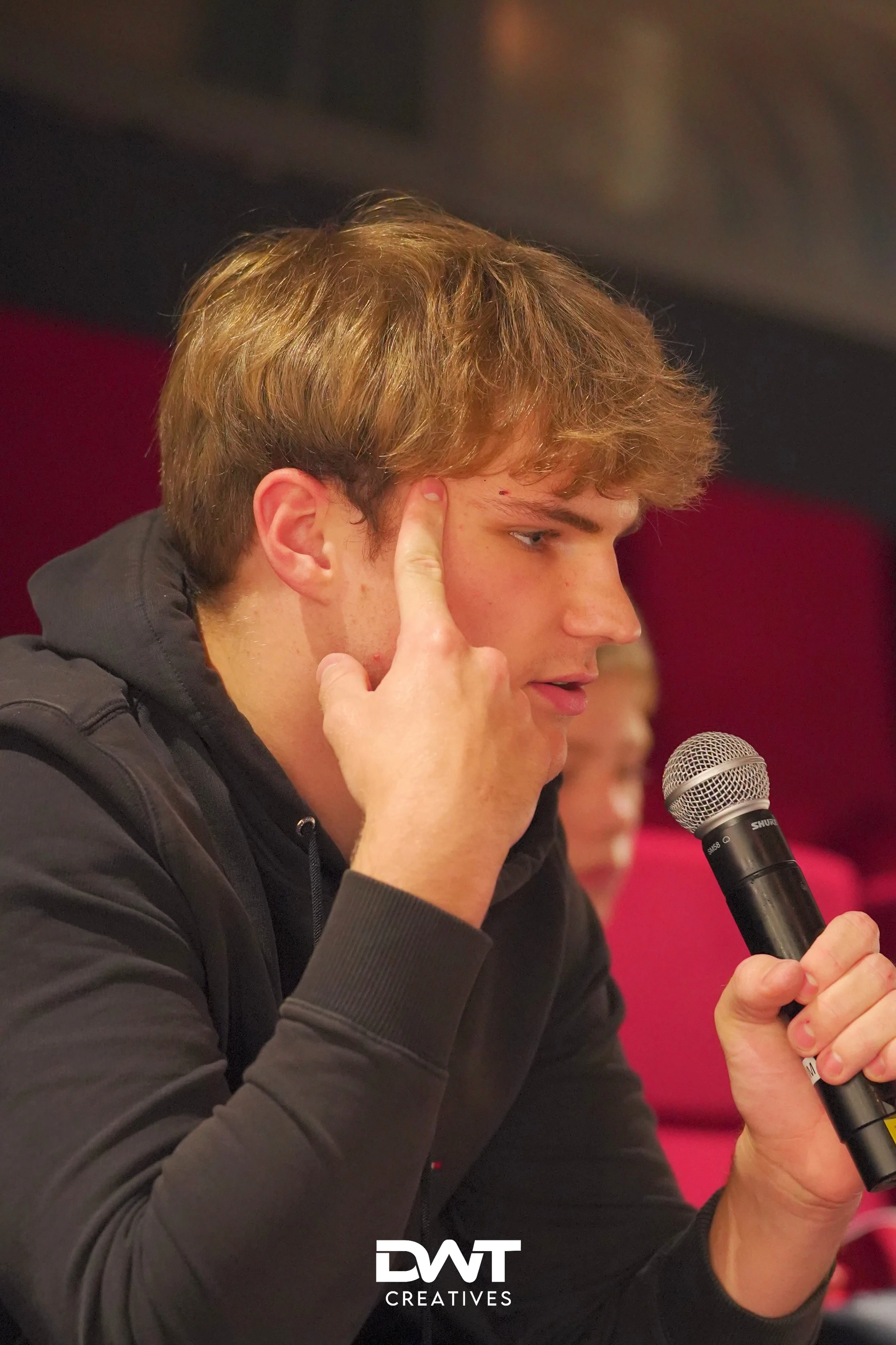 Man met bruine haar in zwarte hoodie praat in een microfoon.