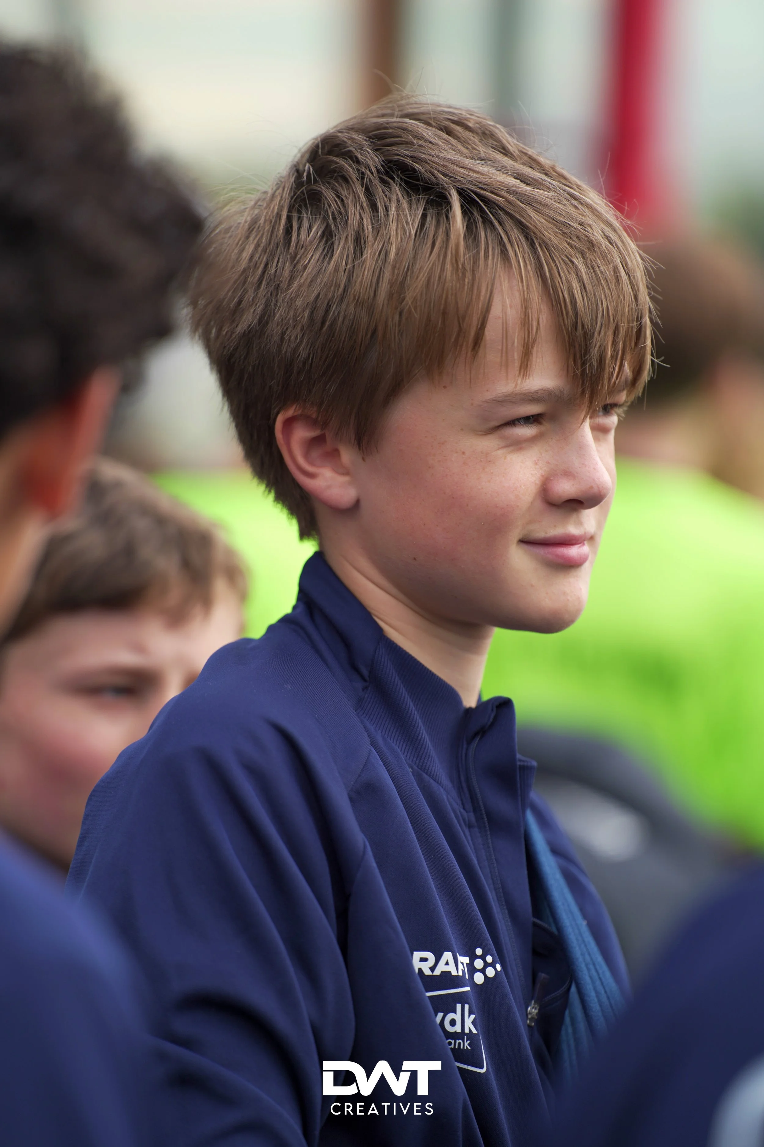 Een jongeman met een donkerblauwe jas glimlacht terwijl hij naar rechts kijkt. Op de achtergrond zijn andere mensen te zien, onscherp in beeld. Het logo van DWT Creatives staat rechtsonder in de foto.