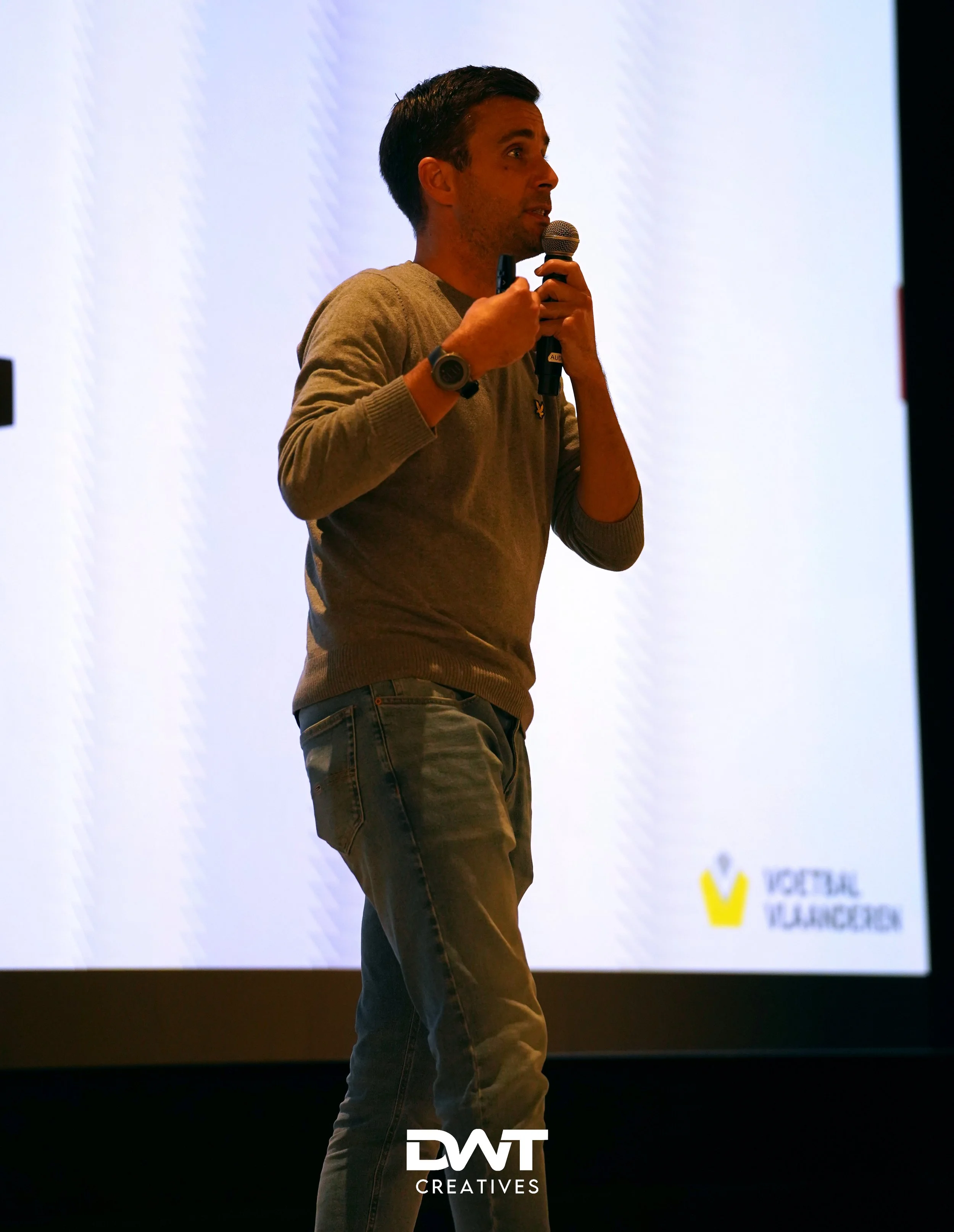 Persoon met microfoon tijdens presentatie op podium met wit scherm en logo "Voetbal Vlaanderen" zichtbaar. Logo "DWT Creatives" op de voorgrond.