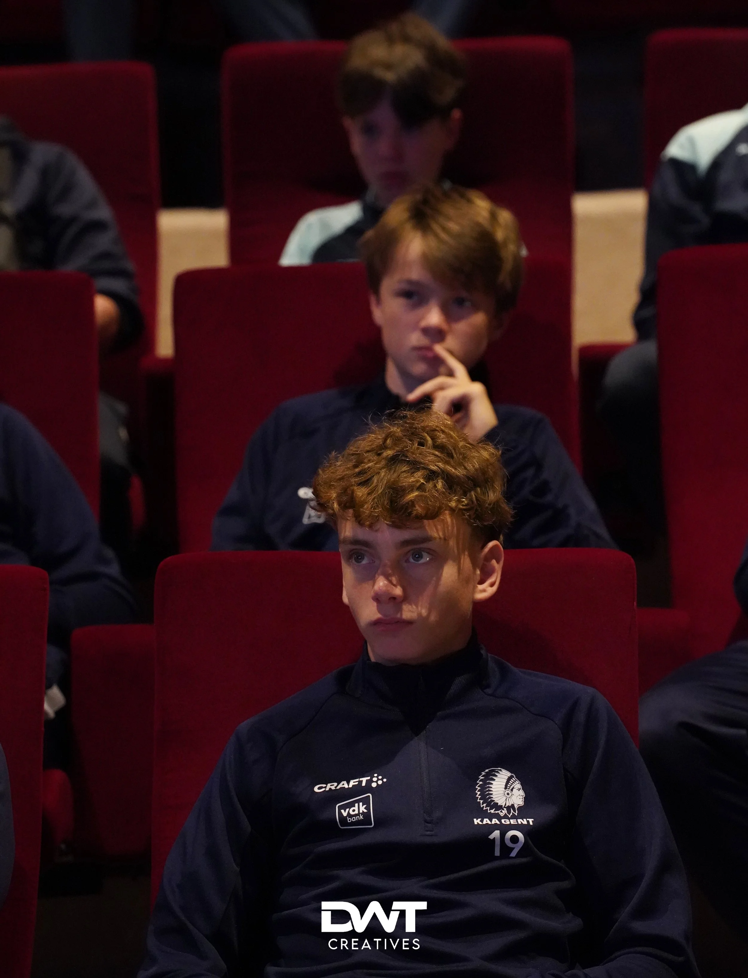 Jongens in sportkleding zitten in een rode theaterstoel, aandachtig kijkend. Het logo van KAA Gent is zichtbaar op hun truien.