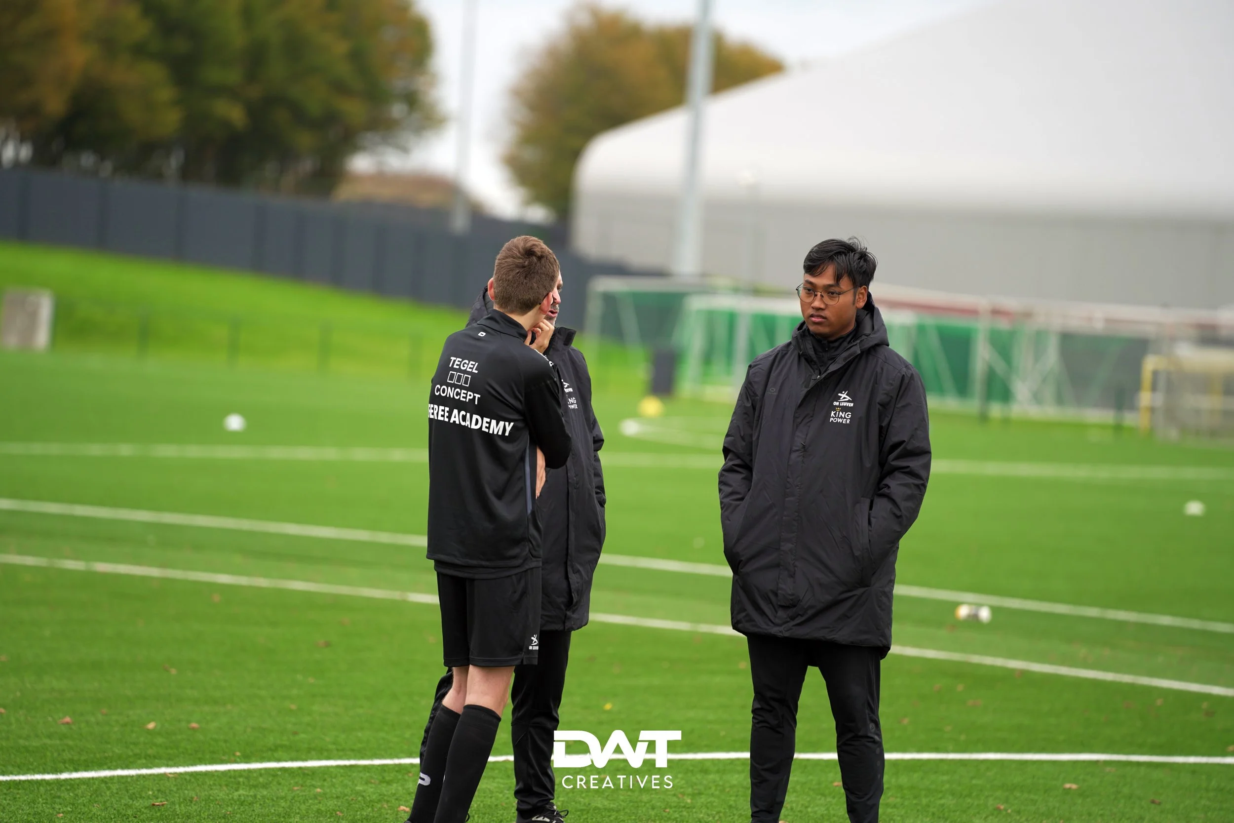 Mensen op kunstgrasveld in sportkleding, mogelijk coaches of trainers, pratend met elkaar, met achtergrond van sportfaciliteiten.