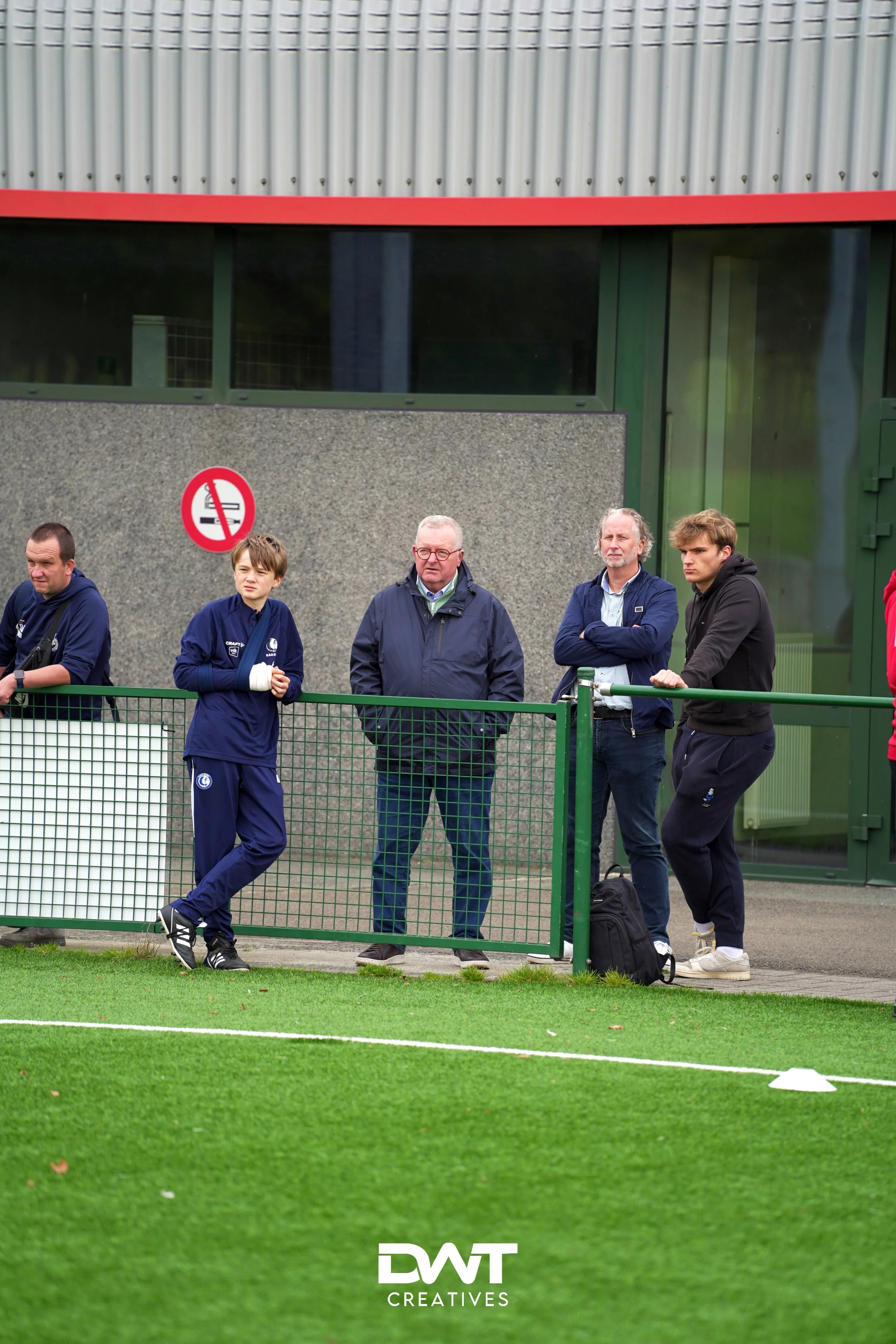 Mensen staan langs een hek bij een sportveld, sommigen leunen tegen het hek, terwijl anderen staan. Er is een bord met rookverbod zichtbaar op een muur op de achtergrond.