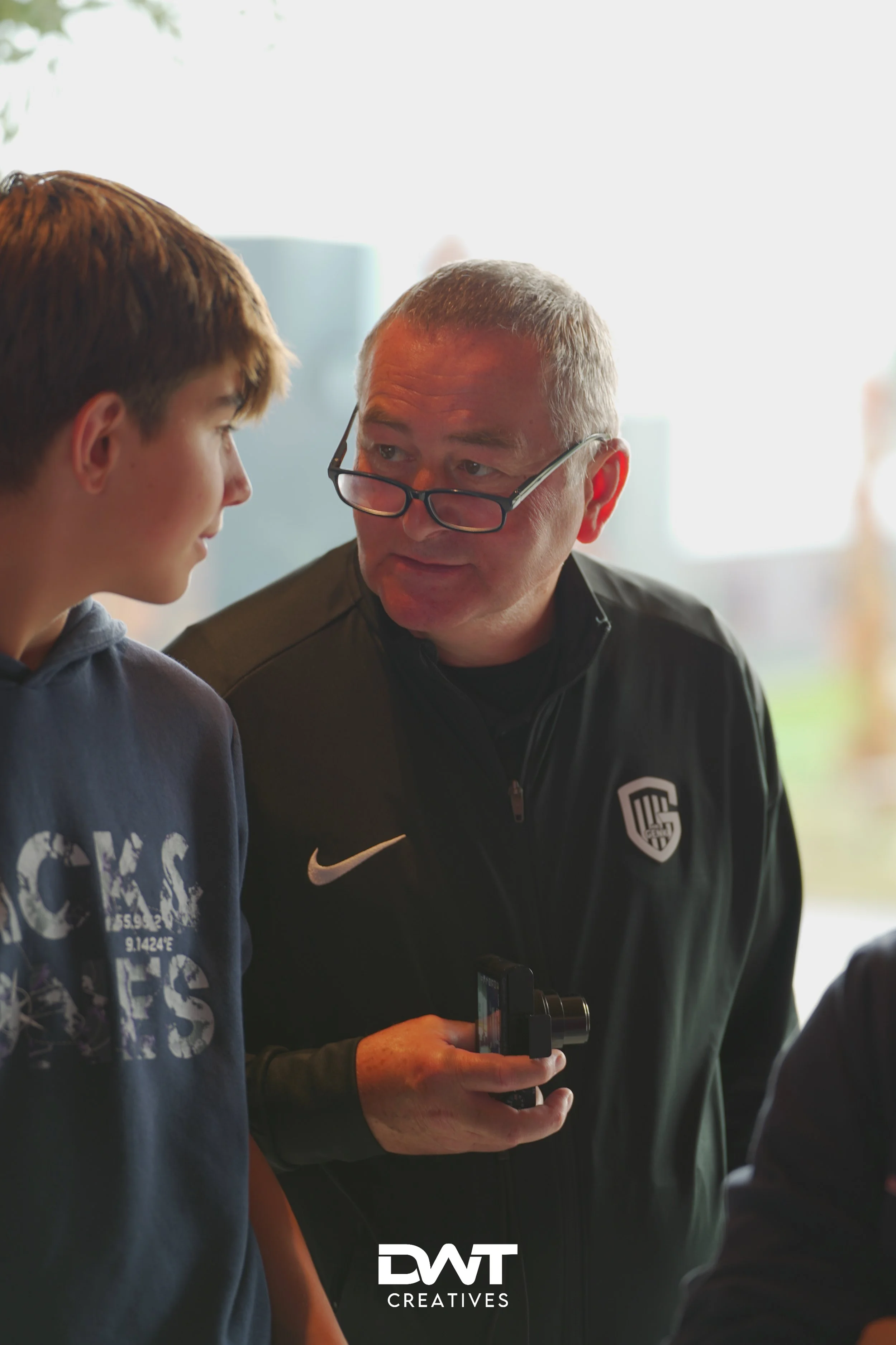Twee mensen in gesprek, een oudere man met bril en een jongere jongen, beide in casual kleding. De man houdt een kleine camera vast. Op achtergrond wazig zicht en DWT Creatives logo onderaan.