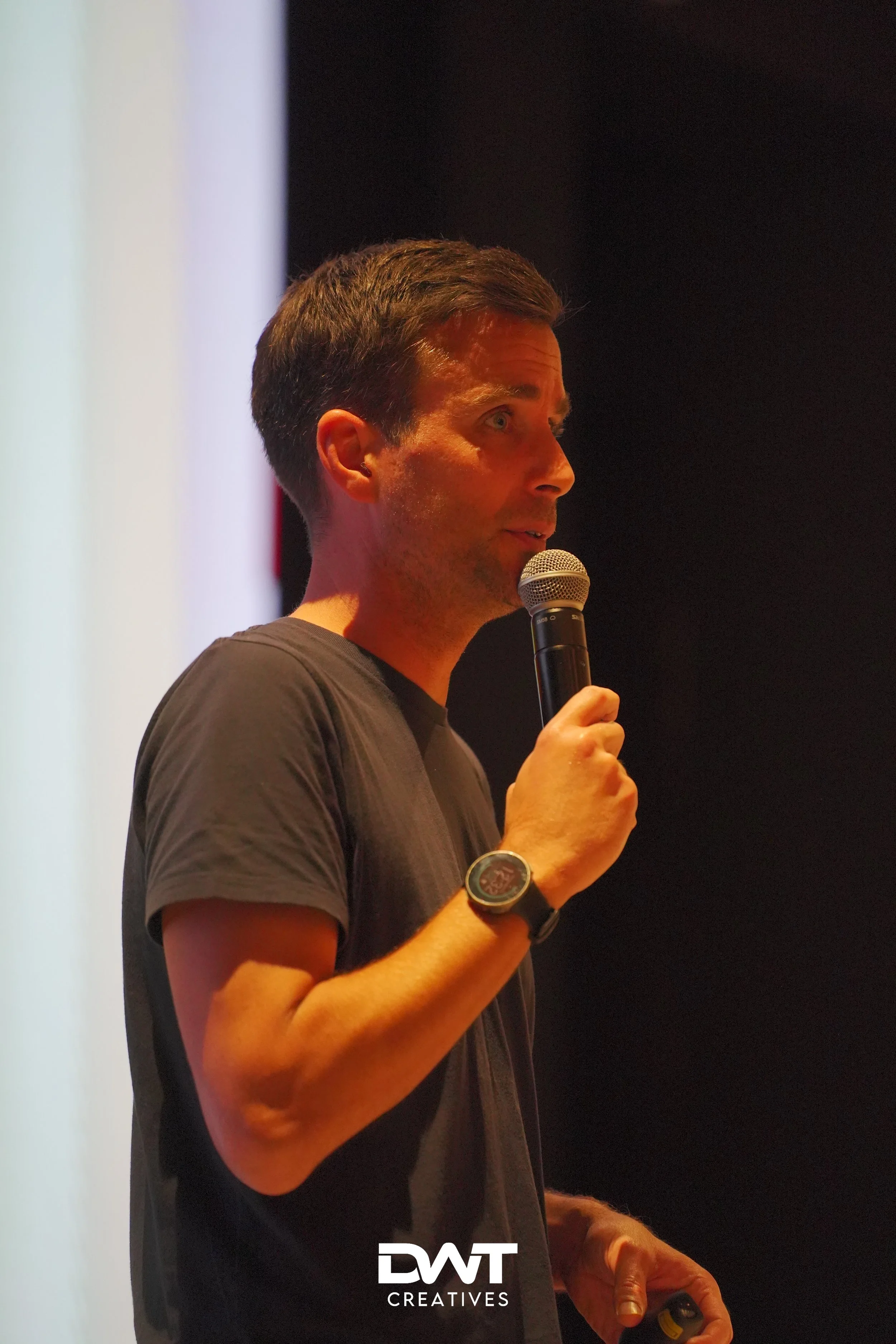 Man met microfoon geeft presentatie op podium, draagt horloge en zwart T-shirt.