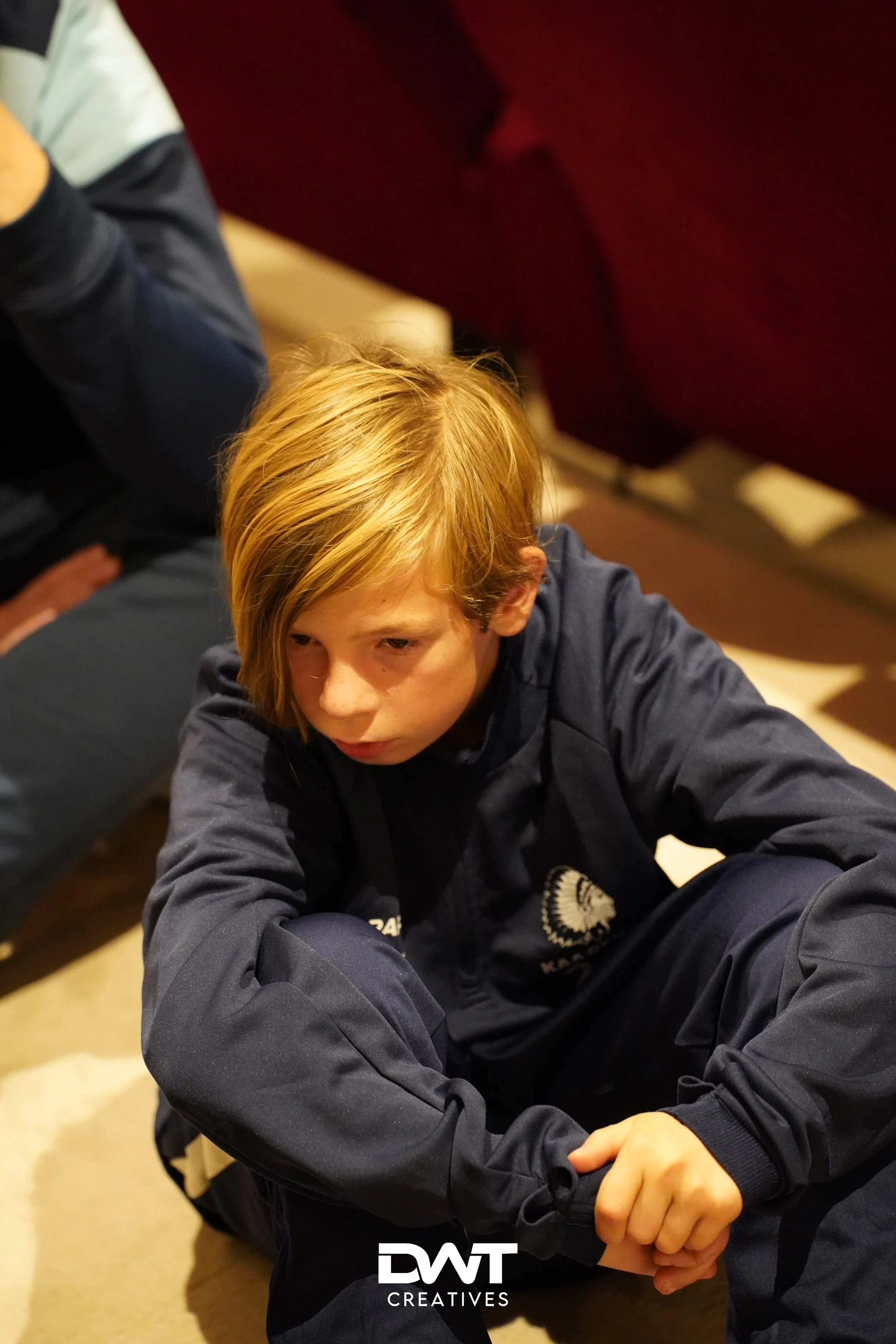 Kind in blauwe sportkleding zit op de grond met gekruiste armen en een nadenkende blik.