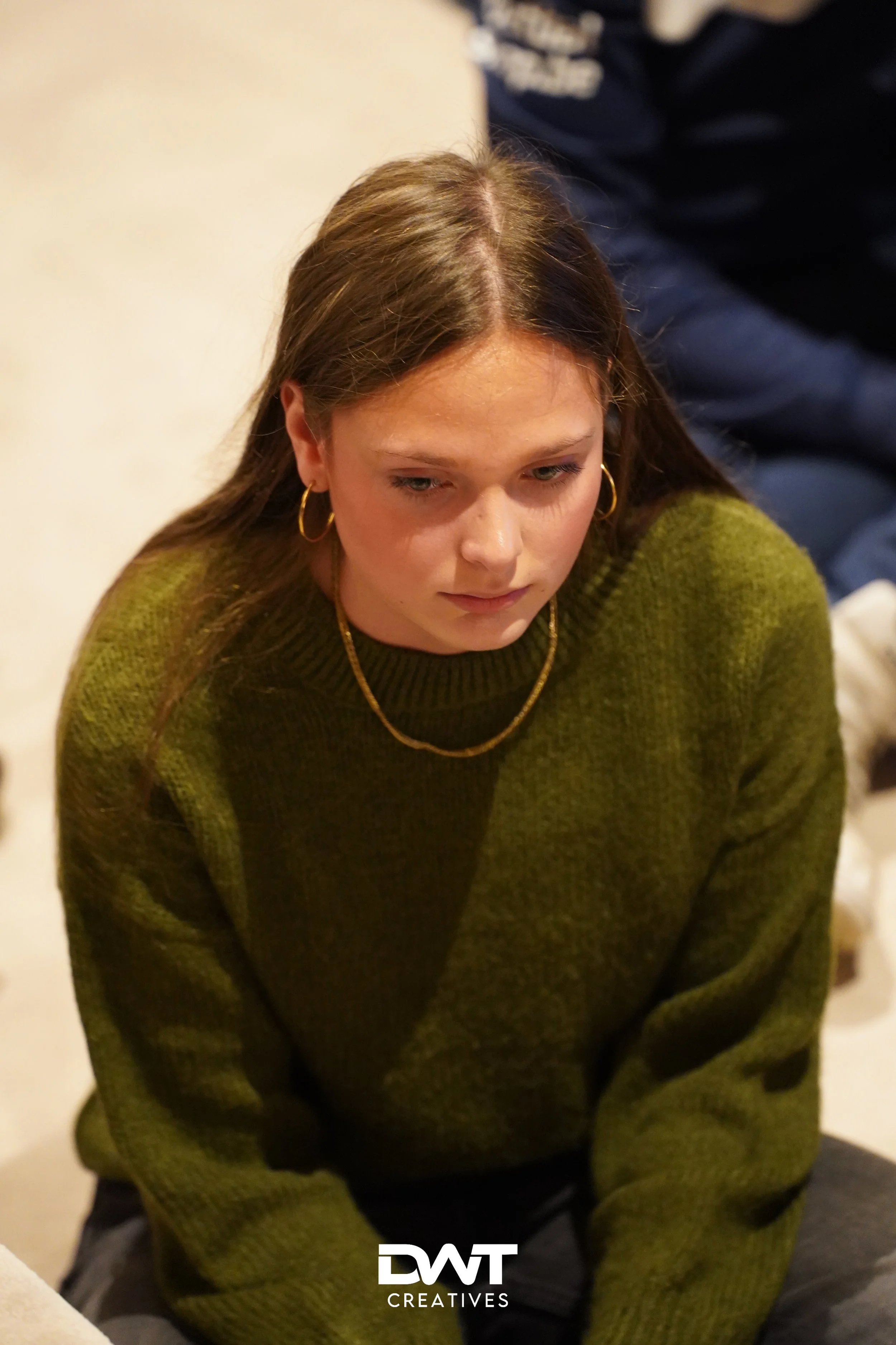 Jonge vrouw met groene trui kijkt naar beneden, zittend op de grond. Ze draagt gouden oorbellen en een ketting.