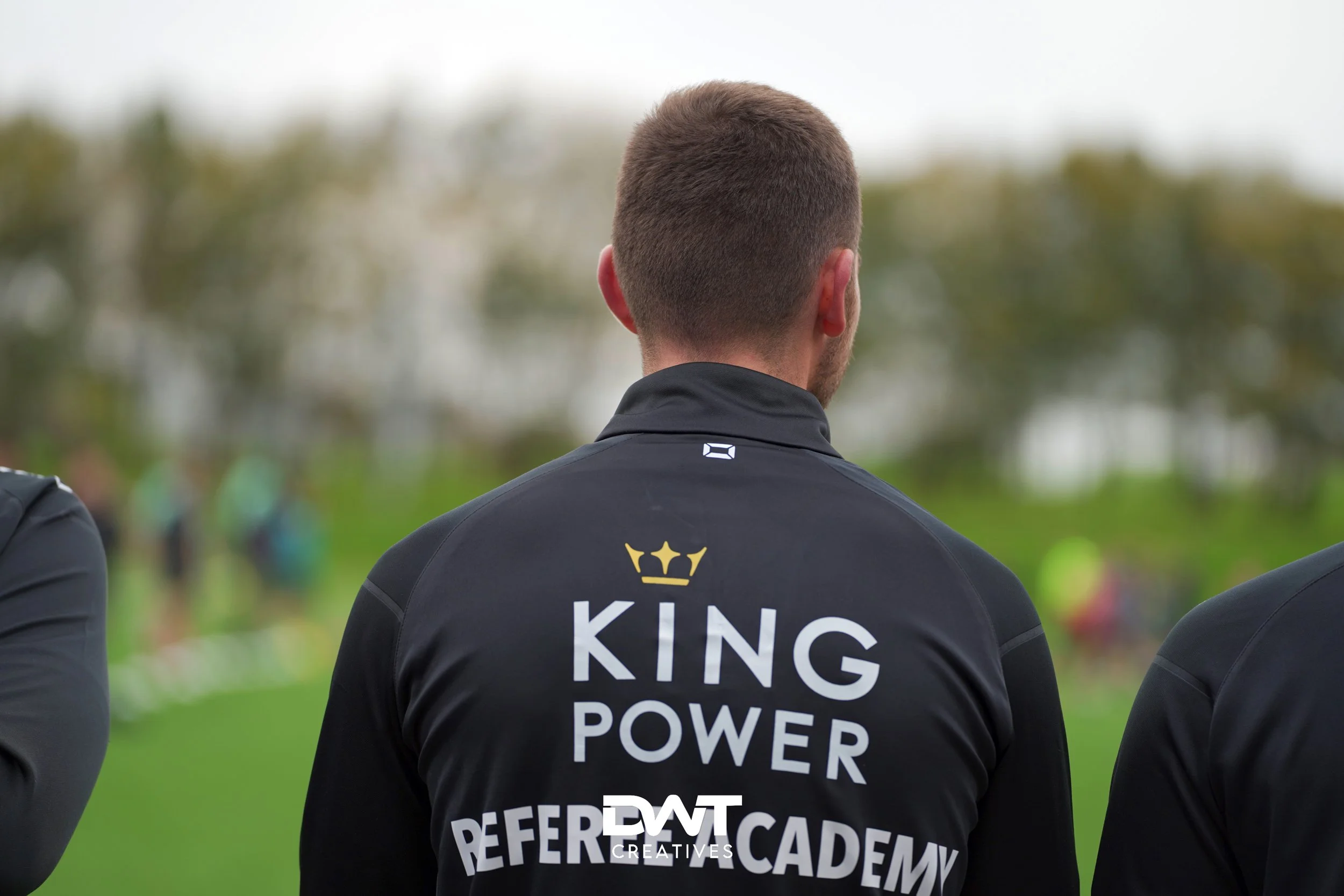 Achteraanzicht van een persoon in een zwarte jas met de tekst "King Power Referee Academy". Personen en bomen zijn vaag op de achtergrond zichtbaar.
