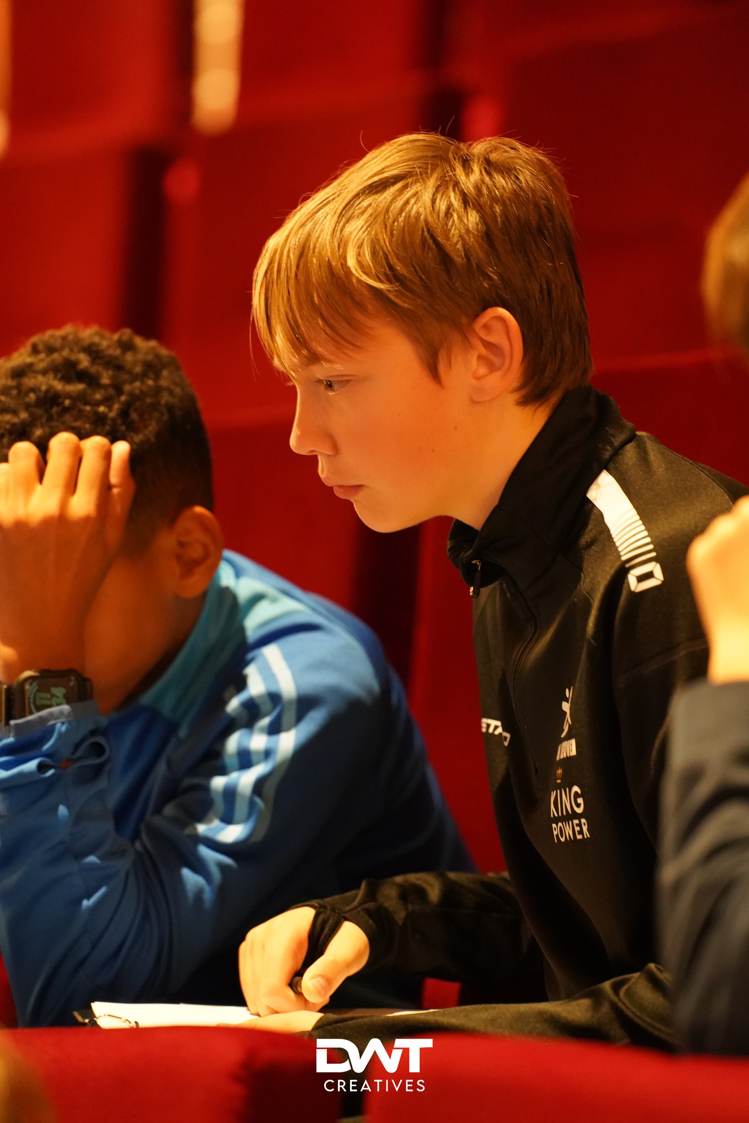 Twee jongens zitten in een rode zaal. De jongen rechts kijkt geconcentreerd en draagt een zwart sportjack. De jongen links ondersteunt zijn hoofd met zijn hand en draagt een blauw sportjack.