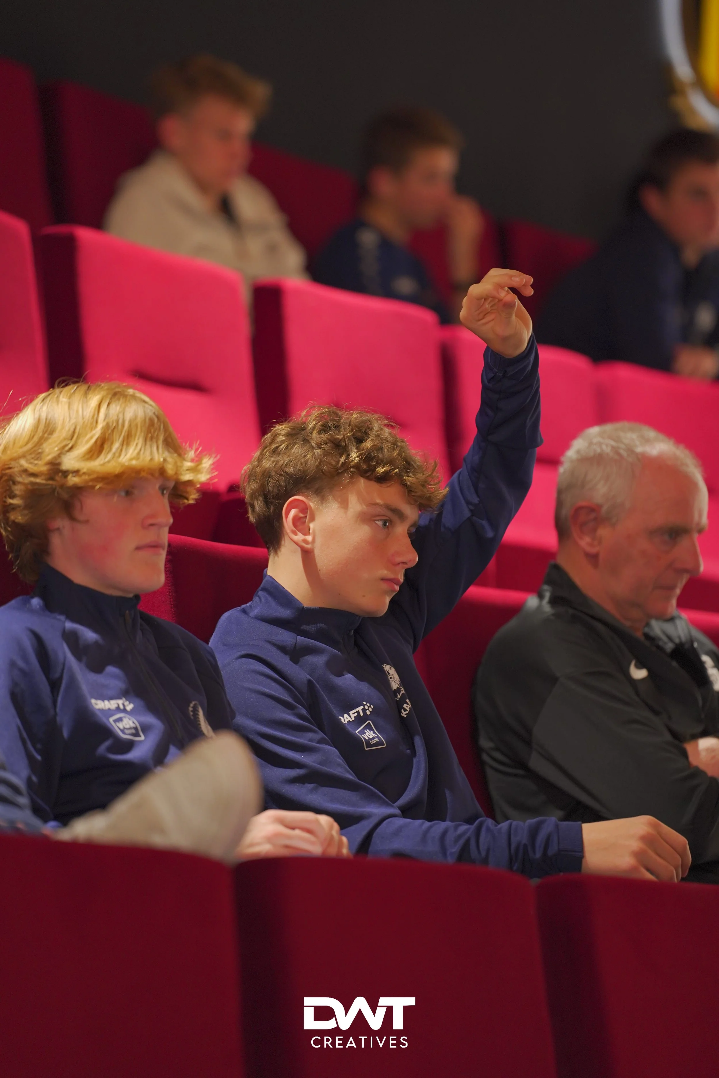 Mensen zitten in een theaterzaal met rode stoelen, één persoon steekt zijn hand op.
