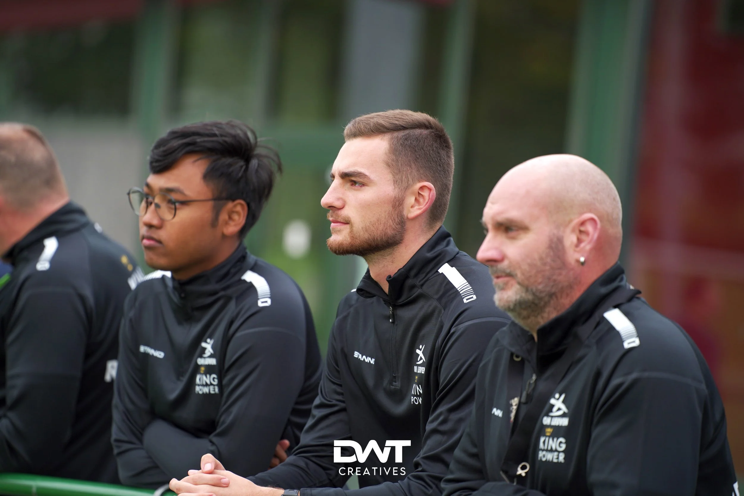 Drie mannen in sportkleding kijken geconcentreerd naar iets buiten beeld. Ze dragen trainingsjacks met opdruk.