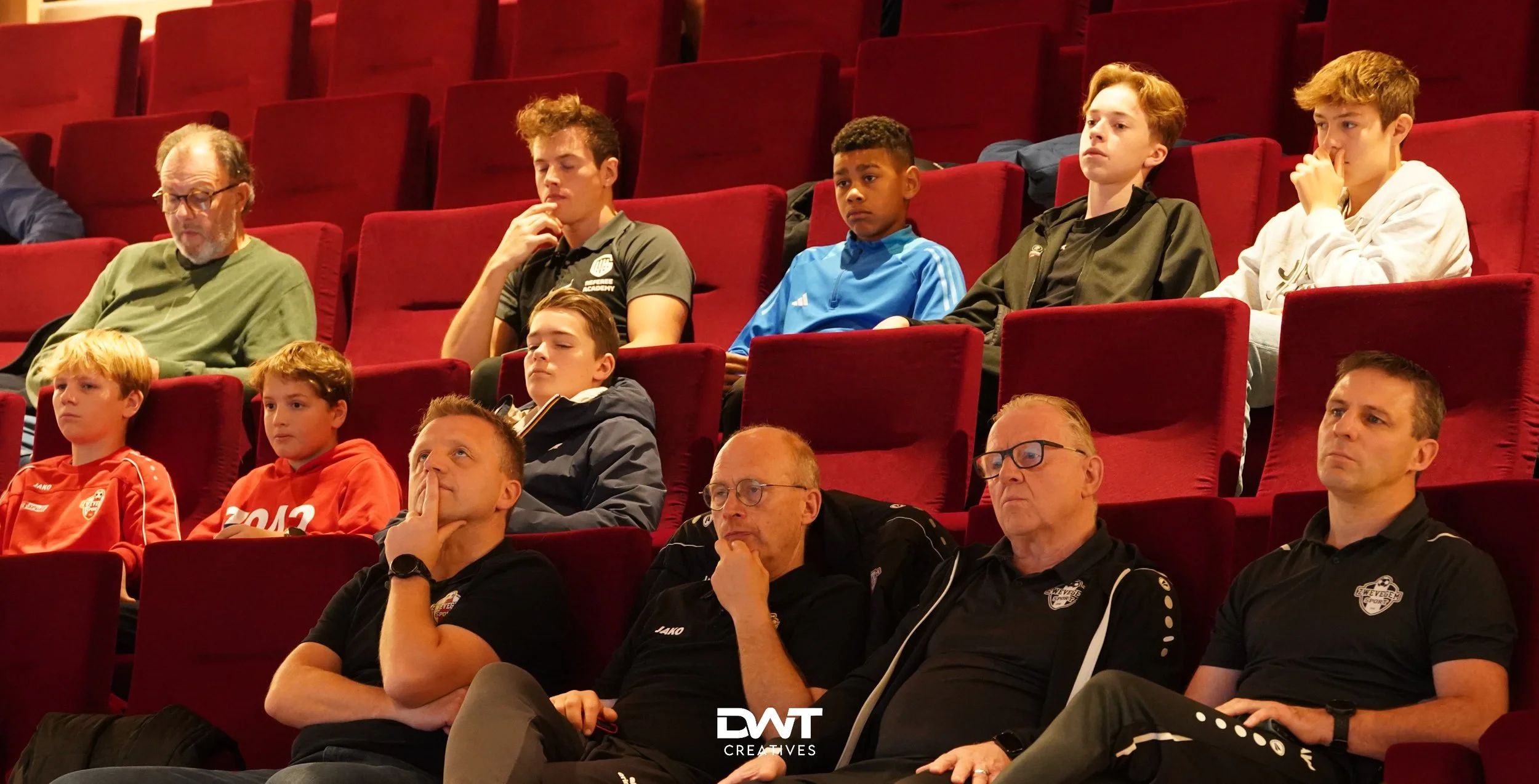 Personen zitten in een theater of bioscoopzaal, aandachtig luisterend, in een rij van bordeauxrode stoelen.