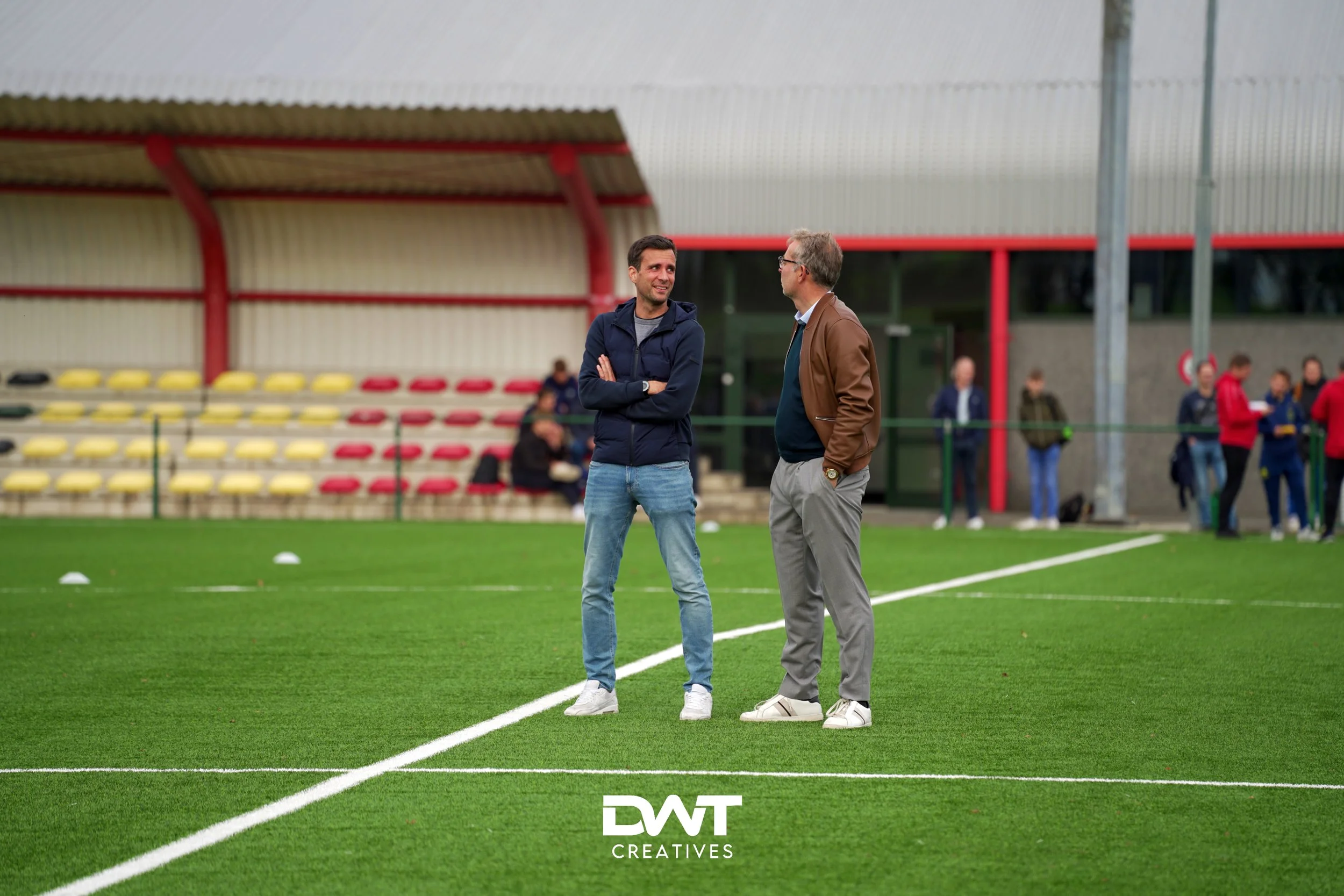 Twee mannen staan op een grasveld voor een tribune met rode en gele zitplaatsen. Ze praten met elkaar. Op de achtergrond staan mensen bij een gebouw met rode accenten. "DWT Creatives" logo is zichtbaar op het grasveld.